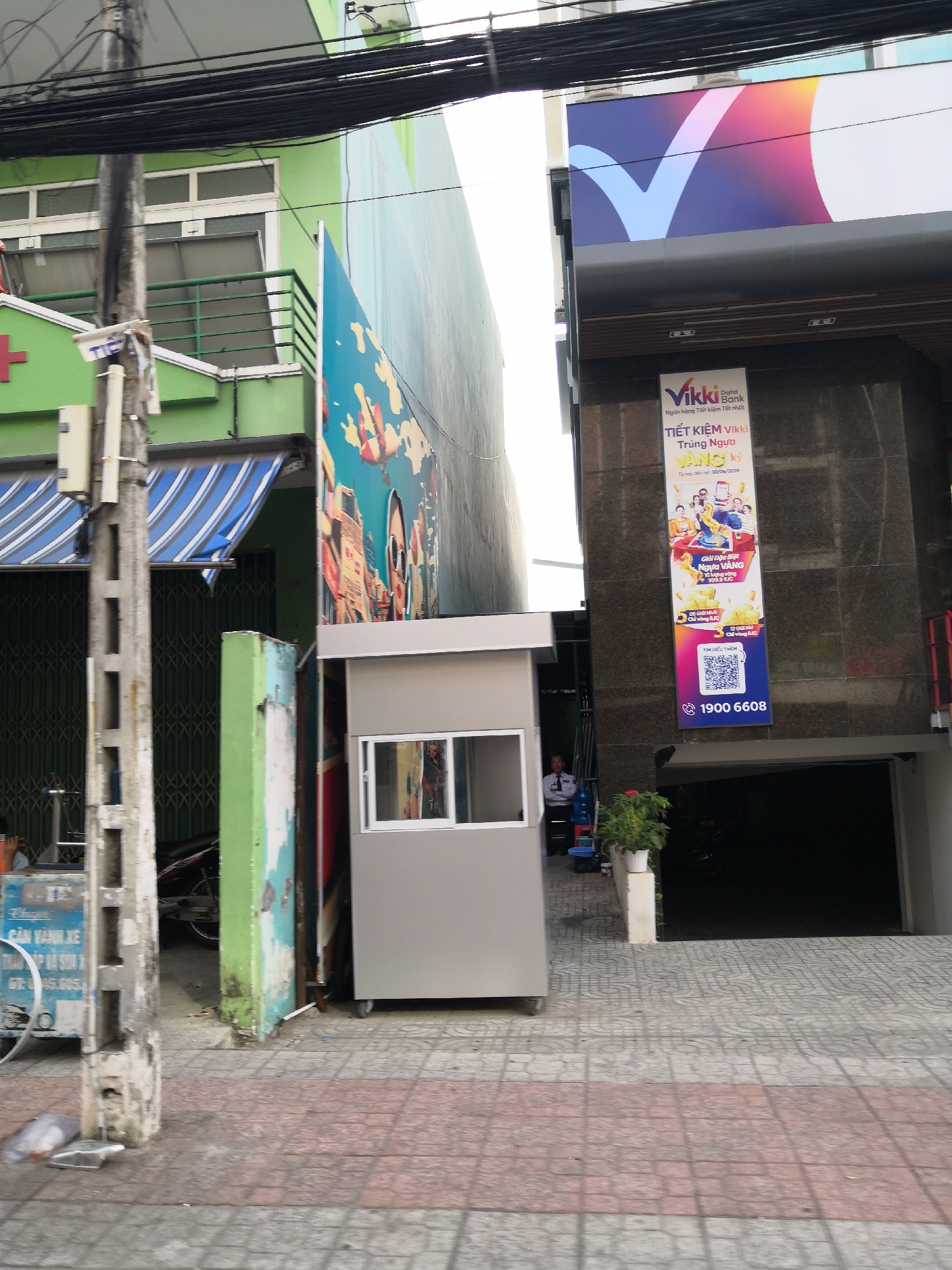 The image depicts a street scene in Nha Trang Ward, Vietnam. In the foreground, a narrow sidewalk with red and grey patterned tiles is visible, with a concrete utility pole on the left. To the right of the pole, a small, grey security booth with a window is situated on the sidewalk. Behind the booth, a dark entryway into a building is present.
Further back, a vibrant mural featuring a person with sunglasses and abstract elements adorns a wall. To the right of the mural, a building facade with a large banner for "Vikki Digital Bank" is prominent. The banner advertises a savings promotion with images of gold and people. Above the bank's banner, a large, colorful logo with a V-shape is displayed on the building's exterior.
To the left of the mural, a green building with striped awnings and a balcony is visible. A red cross symbol is present on this building. Various utility wires are strung overhead, crossing the frame. The overall impression is an urban environment with commercial activity and pedestrian access.