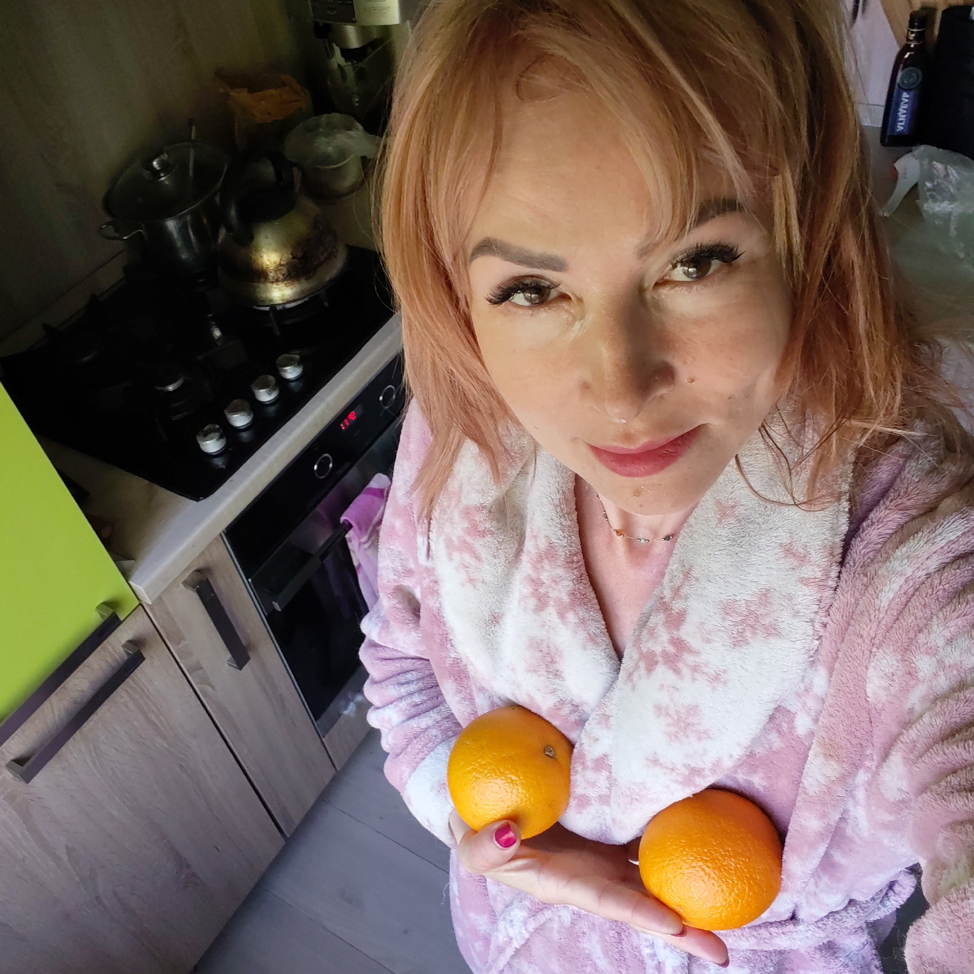 A woman with light pink hair, wearing a pink and white bathrobe and a necklace, is holding two oranges in her hand. She is indoors, standing in a kitchen. Behind her is a black stovetop with a pot and kettle, and kitchen cabinets with light brown wood and green sections. Based on available metadata, the location is in the city of Chaikovsky, Russia.