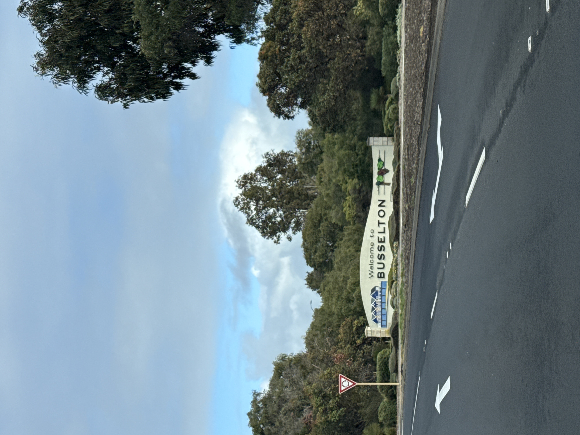 An overhead shot shows a road with white lane markings leading towards a sign that says "Welcome to BUSSELTON." The sign is white with blue and green graphics depicting houses and leaves. Behind the sign, a variety of trees and bushes are visible. The sky above is blue with scattered clouds. A traffic sign depicting a yield ahead is visible to the left of the main sign.