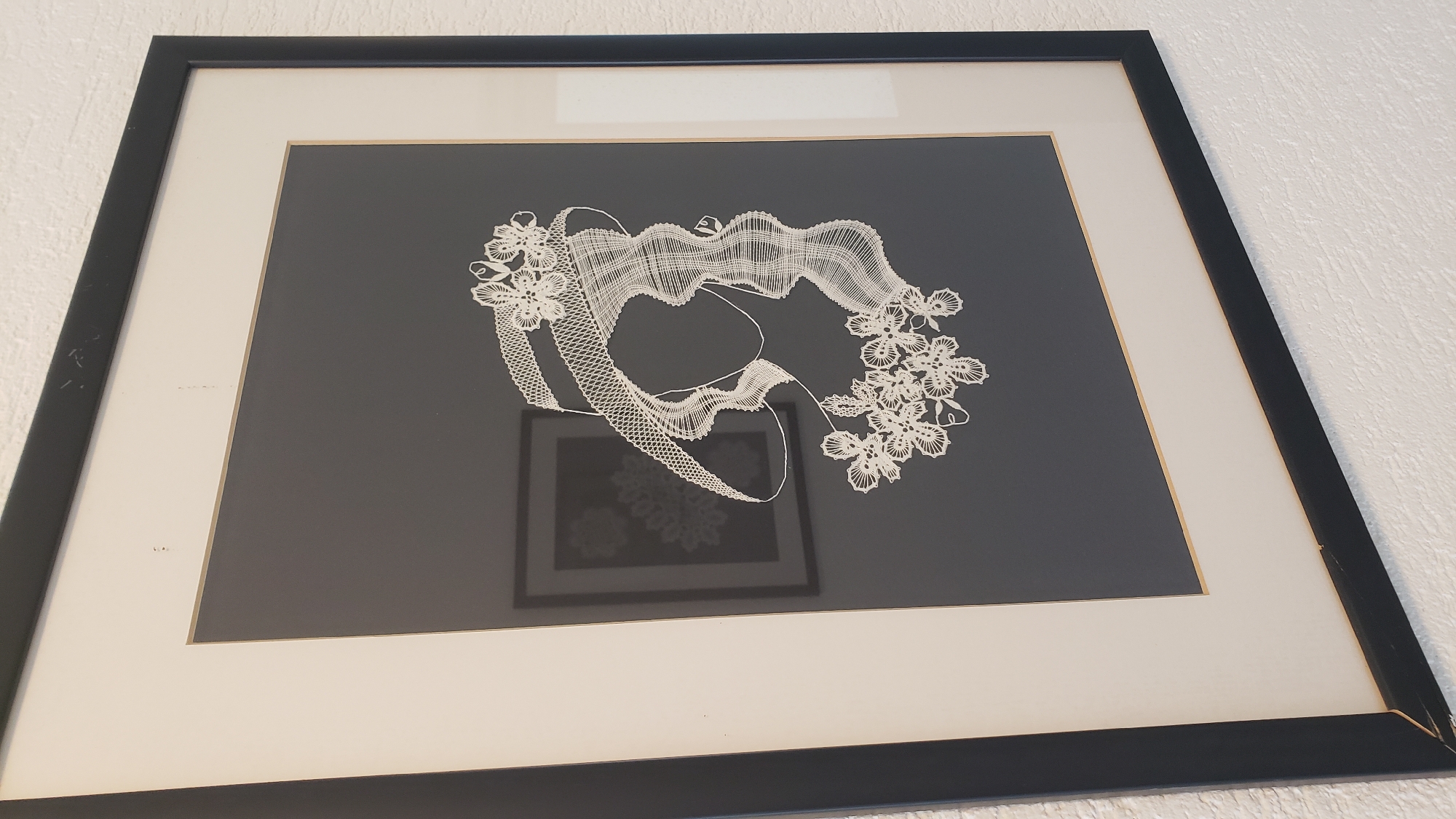 The image shows a framed artwork, a silhouette of a woman's head and shoulders adorned with a hat and flowers, created from intricate white lace. The artwork is matted with a cream-colored border and framed in black. A faint reflection of another framed artwork, also featuring lace patterns, is visible on the glass. The artwork is displayed on a white textured wall. This image was taken in a residential interior in Prešov, Slovakia.