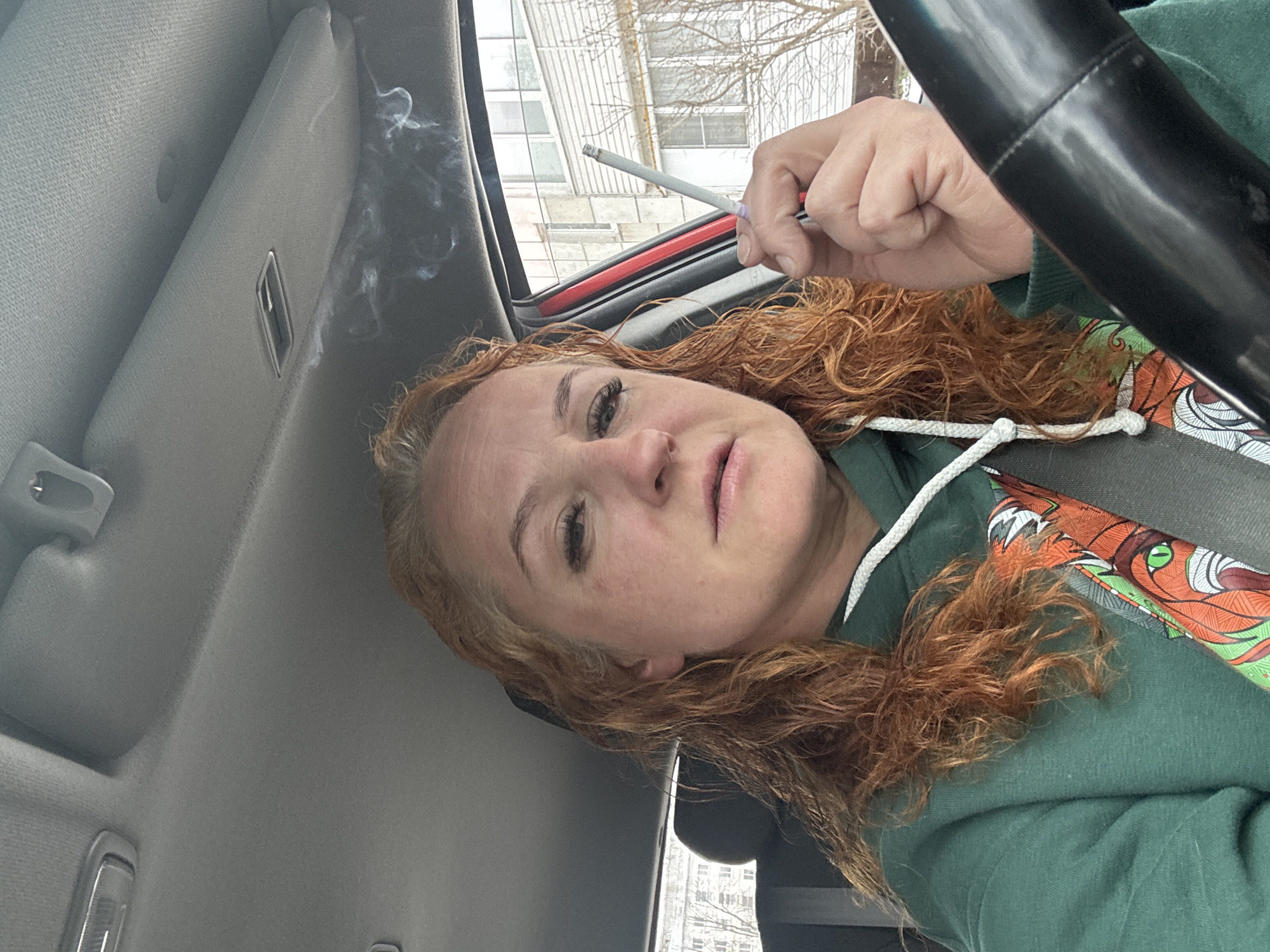 A woman with reddish-blonde curly hair sits in the driver's seat of a car. She is wearing a green hoodie with a graphic design visible on the chest. Her seatbelt is fastened. She holds a lit cigarette in her right hand. Smoke is visible in the car's interior near the ceiling. Outside, a building with multiple windows and bare trees suggest a city environment, identifiable as Chaikovsky, Russia. The perspective is from a slightly low angle, looking upwards toward the woman. A part of the steering wheel is visible in the top right corner of the image.