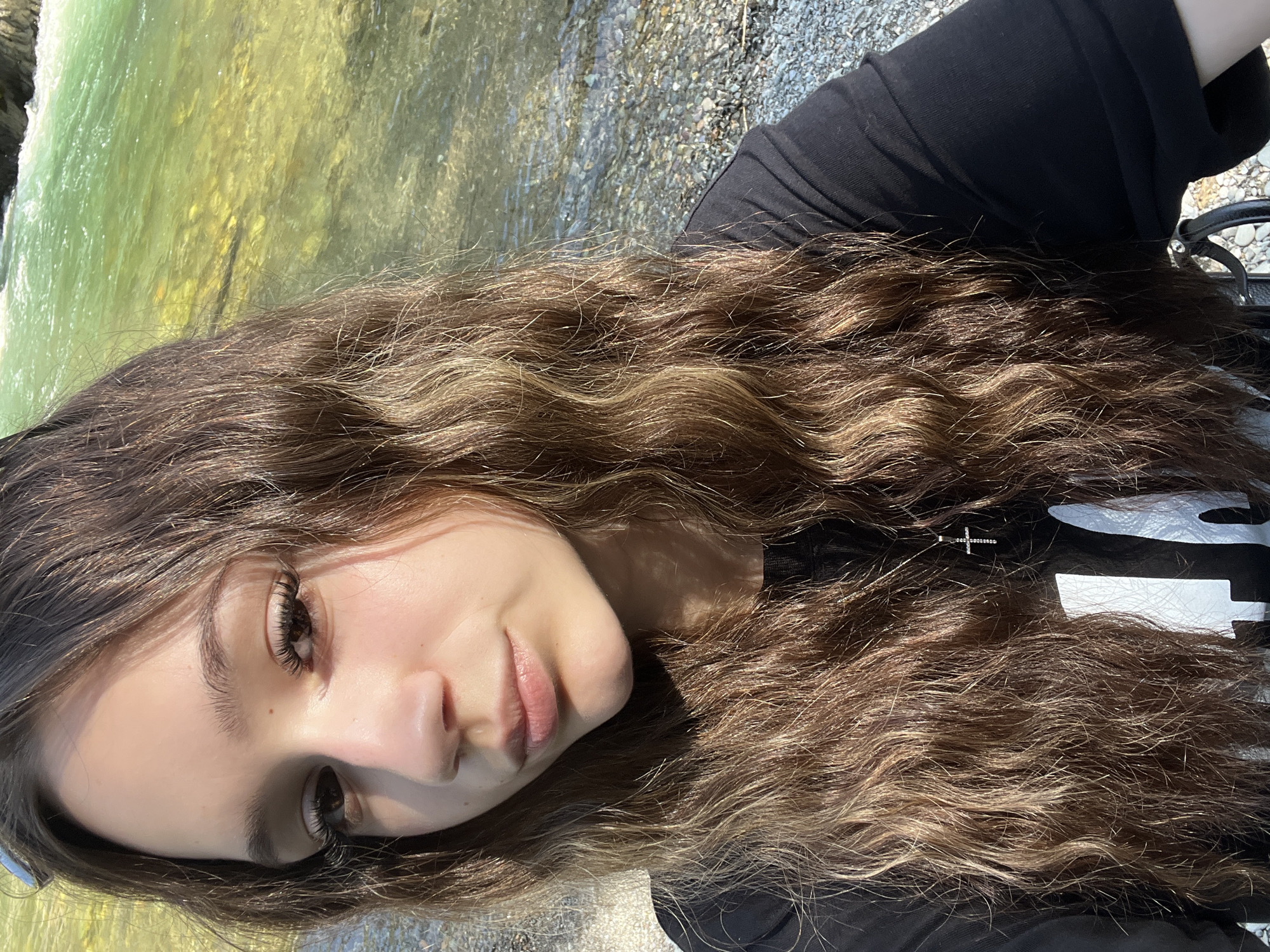 A young woman with long, wavy brown hair is posing for a selfie outdoors. She is wearing a black t-shirt with a white graphic design and a silver cross pendant necklace. Her hair is partially covering her face. She is looking towards the camera with a neutral expression. Behind her, there is a body of clear, shallow water with a rocky bottom and some greenery visible on the far bank, suggesting a natural setting like a river or stream. The lighting indicates it is daytime, possibly late afternoon due to the warm tones and shadows. The background is slightly blurred, emphasizing the subject.