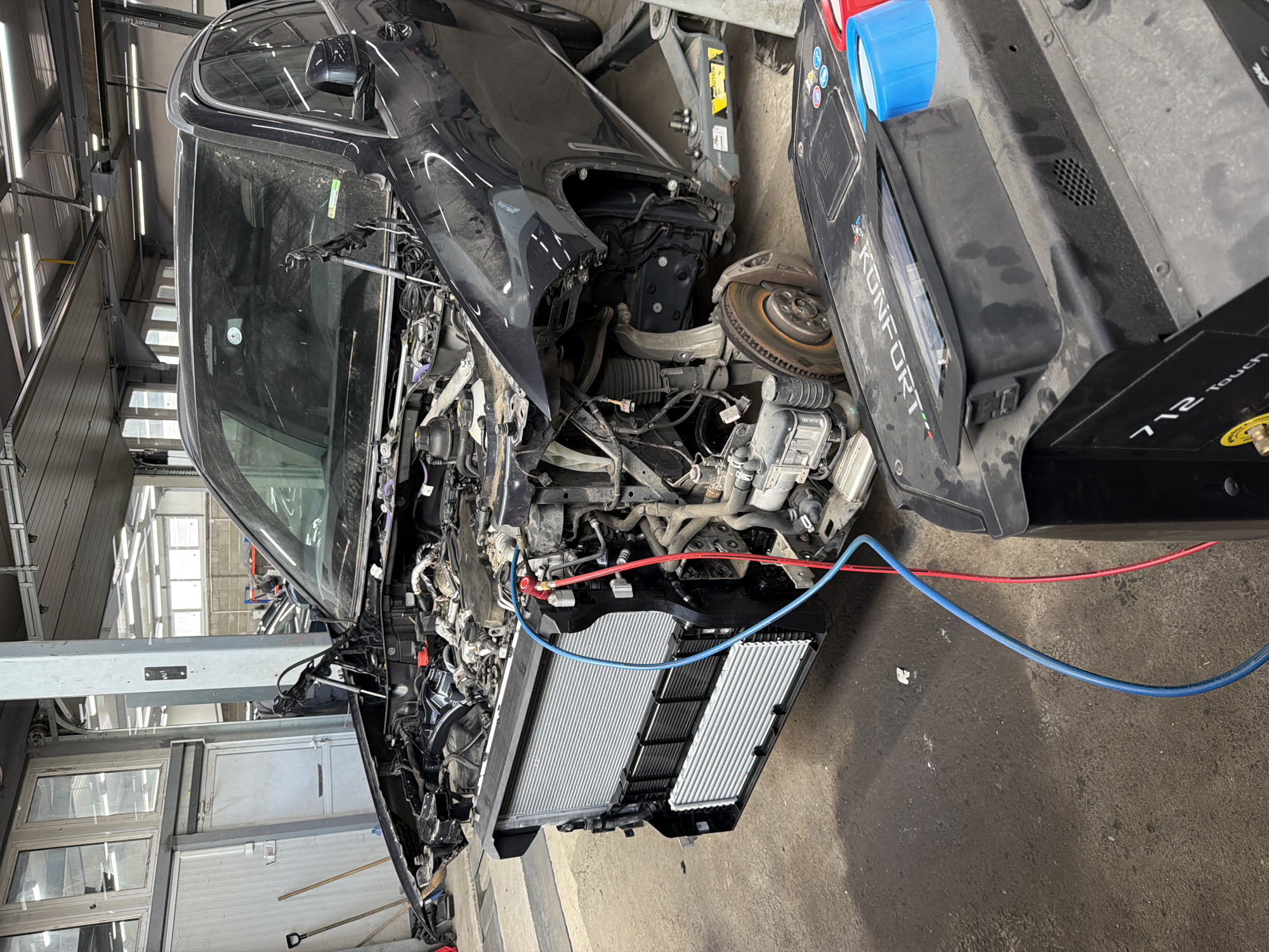 The image shows the front end of a dark-colored car, severely damaged and undergoing repair in what appears to be an auto repair shop. The front panels are dismantled, exposing the engine, radiator, and various mechanical components. The car is positioned on a lift. Red and blue hoses are connected to the car's system, suggesting air conditioning or coolant work.
A Konfort 712 Touch machine is visible next to the car. The environment is a typical garage setting with concrete floors and fluorescent lighting. The location is indicated as Burgas, Bulgaria, based on the provided information, but no specific landmarks or details confirm this location visually.