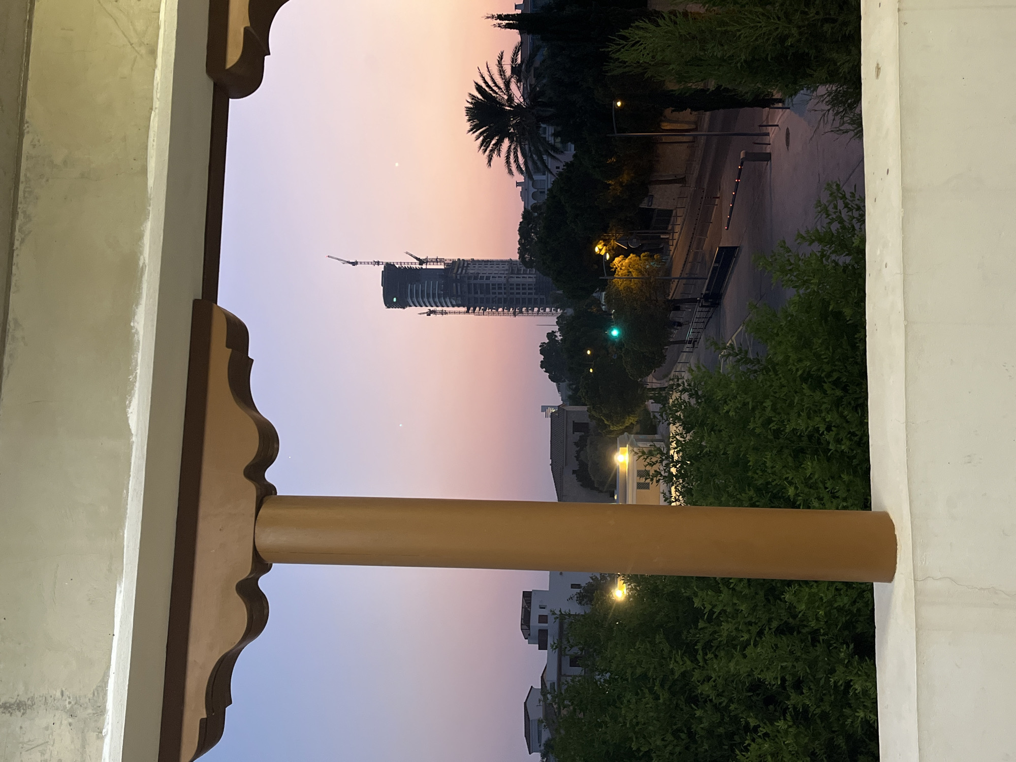 An outdoor, low-angle shot captures a view through an ornate, dark brown wooden archway onto a street scene at dusk in Limassol, Cyprus. The sky is a soft gradient of lavender and pink, with two faint stars visible. In the mid-ground, a tall, unfinished skyscraper with visible scaffolding and cranes dominates the skyline. To its right, a palm tree stands prominently. A street with streetlights and a traffic barrier with red lights is visible, bordered by trees and buildings. A traffic light shows a green signal. In the foreground, a thick, brown wooden pillar on the left and a white concrete ledge frame the view. Lush green foliage is abundant throughout the scene, with some trees appearing to be cypress. The overall impression is of a city undergoing development, observed during the tranquil transition from day to night.