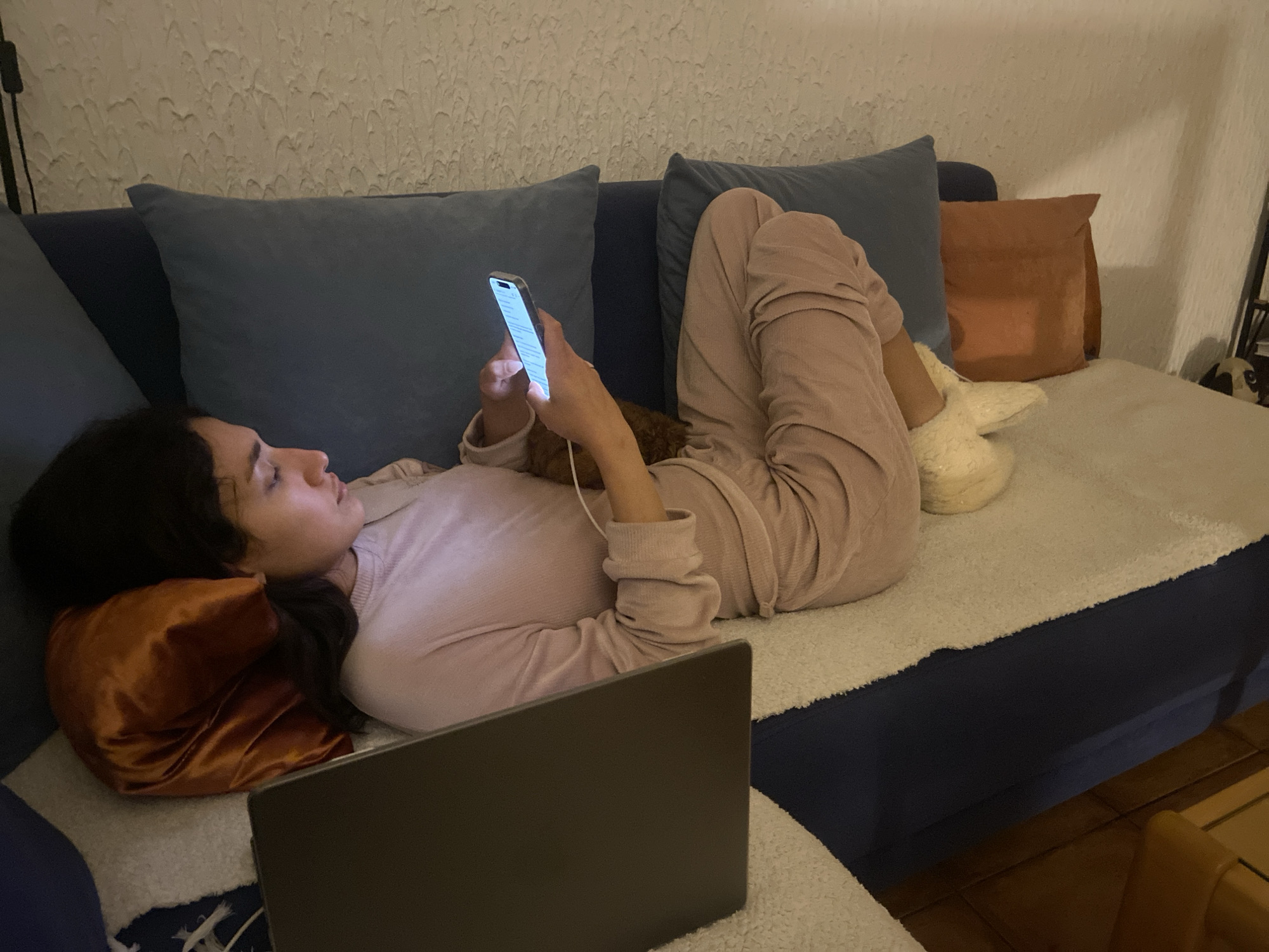 A woman is lying on a blue couch in an interior space, likely a home. She is wearing a light pink outfit and white slippers. She is holding a lit smartphone in her right hand and appears to be looking at it. A laptop is in the foreground, partially obscuring the view. There are several pillows on the couch: two are blue, one is brown, and another is orange. The wall behind the couch is textured. The location is not discernible from the image and cannot be identified as Sutomore, Montenegro.