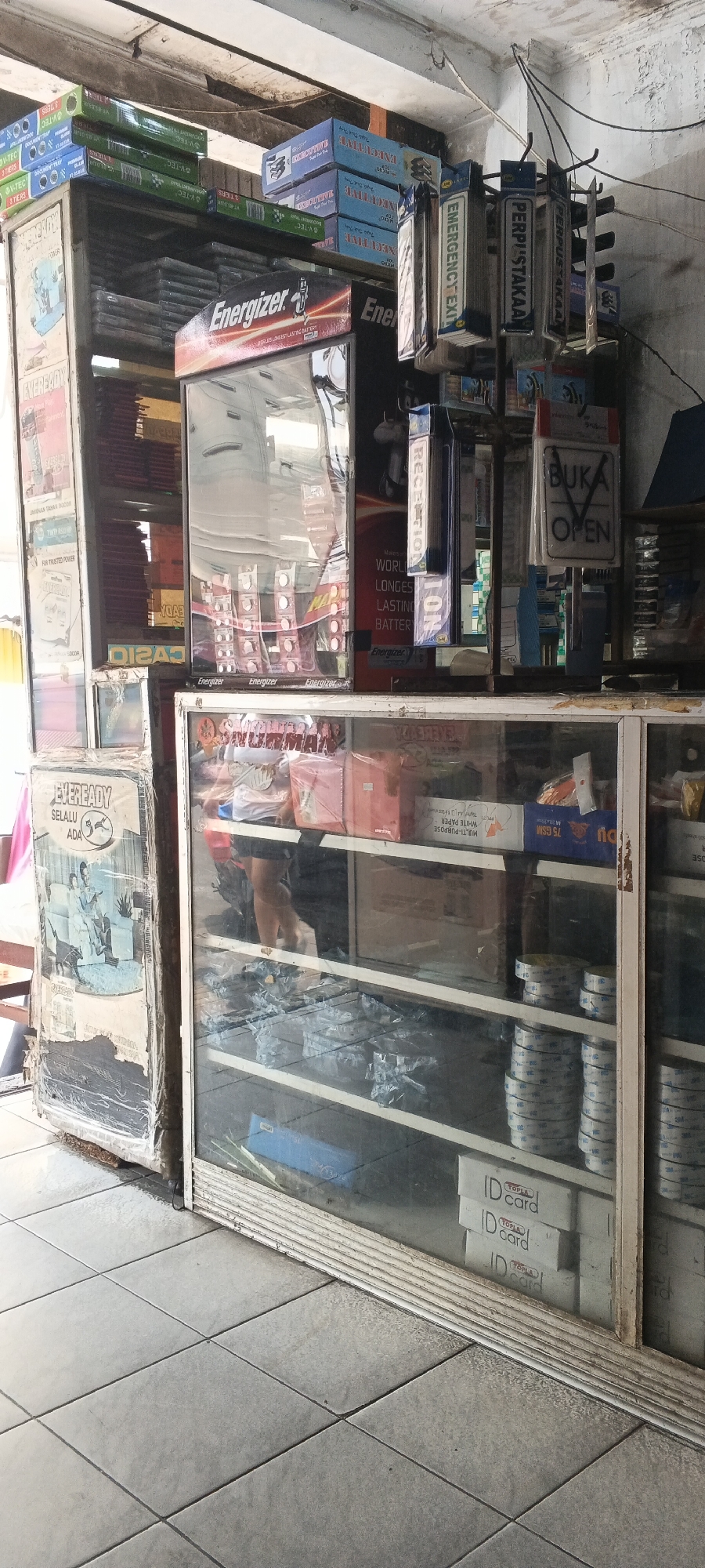The image shows the interior of a small shop, likely a convenience store or a general store, located in Tangerang, Indonesia. The shop appears to be cluttered with various merchandise. In the foreground, a glass display case is visible, filled with stacked items such as rolls of tape and boxes of "ID card" supplies. Above this display, another shelf holds more boxes and items, including what appears to be printer paper.
To the left of the display case, a large, somewhat damaged advertisement poster for "Eveready" batteries is affixed to a tall rectangular structure. This structure might be a refrigerator or another type of display unit. Above the Eveready poster, several shelves are packed with boxes, many of them green and blue, possibly containing stationery or electronic components.
Dominating the center of the image is a refrigerated display case for "Energizer" batteries, with its illuminated signage clearly visible. Behind this, more shelves are stocked with merchandise, and several items are hanging from a rack, including signs that read "EMERGENCY EXIT" and "PERPUSTAKAAN" (which means library in Indonesian). The overall environment suggests a busy retail space with limited organization. The flooring consists of square grey tiles. The lighting is likely artificial, contributing to a somewhat dim and busy atmosphere.
