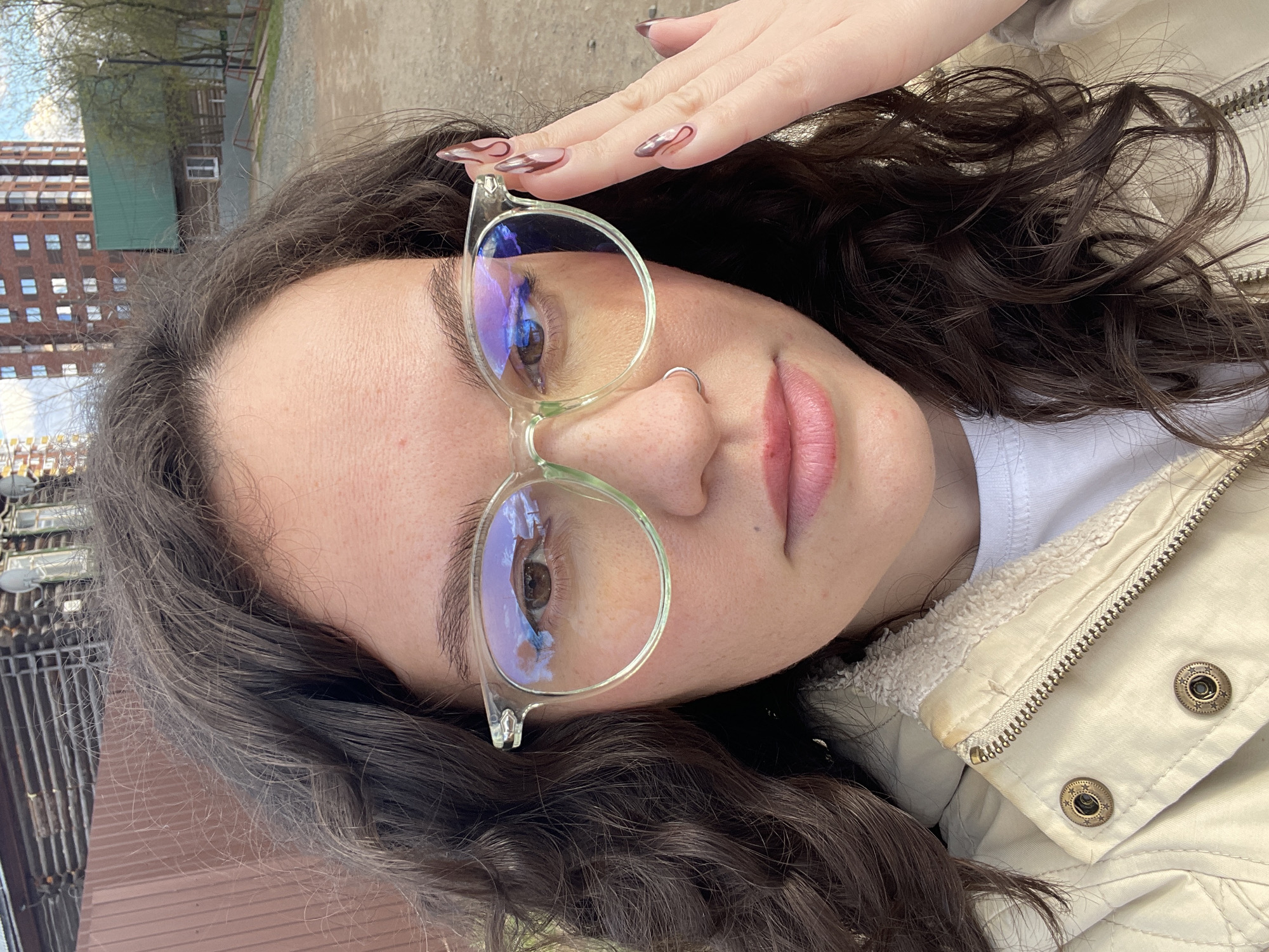 A close-up, slightly tilted portrait captures a person, likely female, with dark, curly hair and light skin, looking directly at the camera. The subject is wearing modern, clear-framed eyeglasses, which reflect a blurred blue and purple light. One hand is raised to the side of the head, revealing long fingernails adorned with an intricate brown and pink swirly design.
The person has pink-toned lips and a small silver hoop nose ring. A light-colored (beige or cream) jacket with a visible zipper and antique-style metal snap buttons featuring a star design is worn over a white t-shirt, with a soft, textured lining visible around the collar.
Reflections in the eyeglasses and visible background elements show an outdoor setting during daytime, featuring green trees and buildings, including multi-story brick structures and a building with a green roof. This urban or suburban environment is consistent with the provided location context of Izhevsk, Russia. The lighting is bright and natural. No visible text is present in the image.