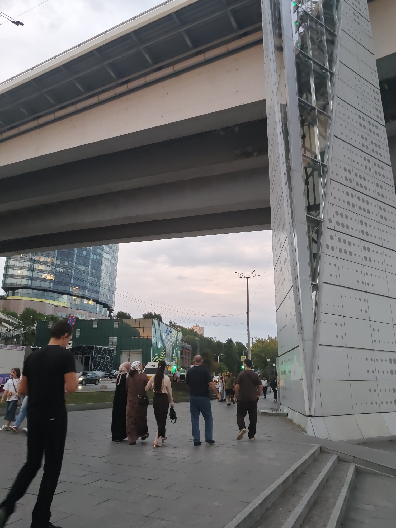 The image shows a modern architectural structure on the right, which appears to be part of a pedestrian overpass or a building facade, with a reflective glass enclosure running vertically through it. This structure is white and decorated with a pattern of gray circles of varying sizes. To the left, a large elevated road or bridge casts a shadow over the scene. In the background, a modern glass-fronted building is visible, along with some trees and other structures.
Several people are present on a paved walkway in the foreground and midground. On the left, a man in a black t-shirt and black pants is walking away from the camera. To his right, a group of four people, three women and a man, are walking together. Two of the women are wearing hijabs and long dresses, and another woman is wearing a beige top and a dark skirt. A man in a dark t-shirt and dark pants is walking behind this group. Further to the right, a man in a blue shirt and blue jeans is walking towards the right edge of the frame. Other individuals and groups are visible in the background, including a person in a white t-shirt and jeans, and another man in a dark t-shirt.
The ground is paved with large, dark gray square tiles. Steps are visible on the right side of the frame leading down from the walkway. A street light pole with a circular fixture is visible in the mid-background. The sky is cloudy.
The architectural style and the presence of the city's infrastructure suggest an urban environment. The specific location is the area around the "Rostov Arena" stadium in Rostov-on-Don, Russia, specifically near the pedestrian connection leading to the stadium, often referred to as the "Rostov Arena Bridge" or the area adjacent to the stadium's elevated pedestrian walkways and modern architectural elements. This area is known for its contemporary design and integration with public spaces.