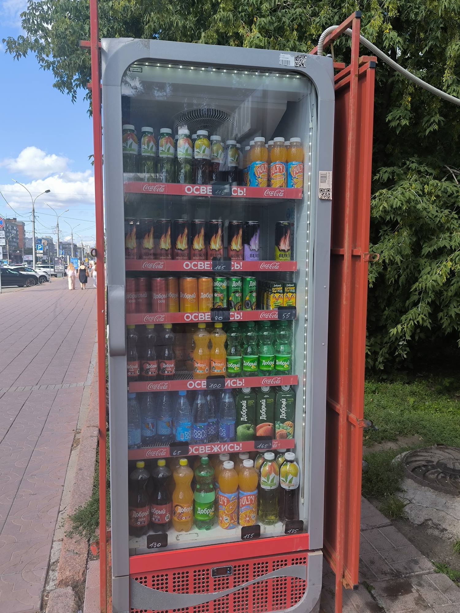 A glass-fronted vending machine is situated outdoors on a sidewalk in Novosibirsk, Russia. The machine is filled with various beverages, including bottled water, soda cans, and juice bottles. Several price tags are visible on the shelves. In the background, trees and a cityscape with buildings and a street can be seen. The sidewalk is paved with red bricks, and there is grass and a manhole cover next to the machine. The sky is blue with some clouds. There are a few people walking on the sidewalk in the distance. The vending machine has a red frame and a grey front.