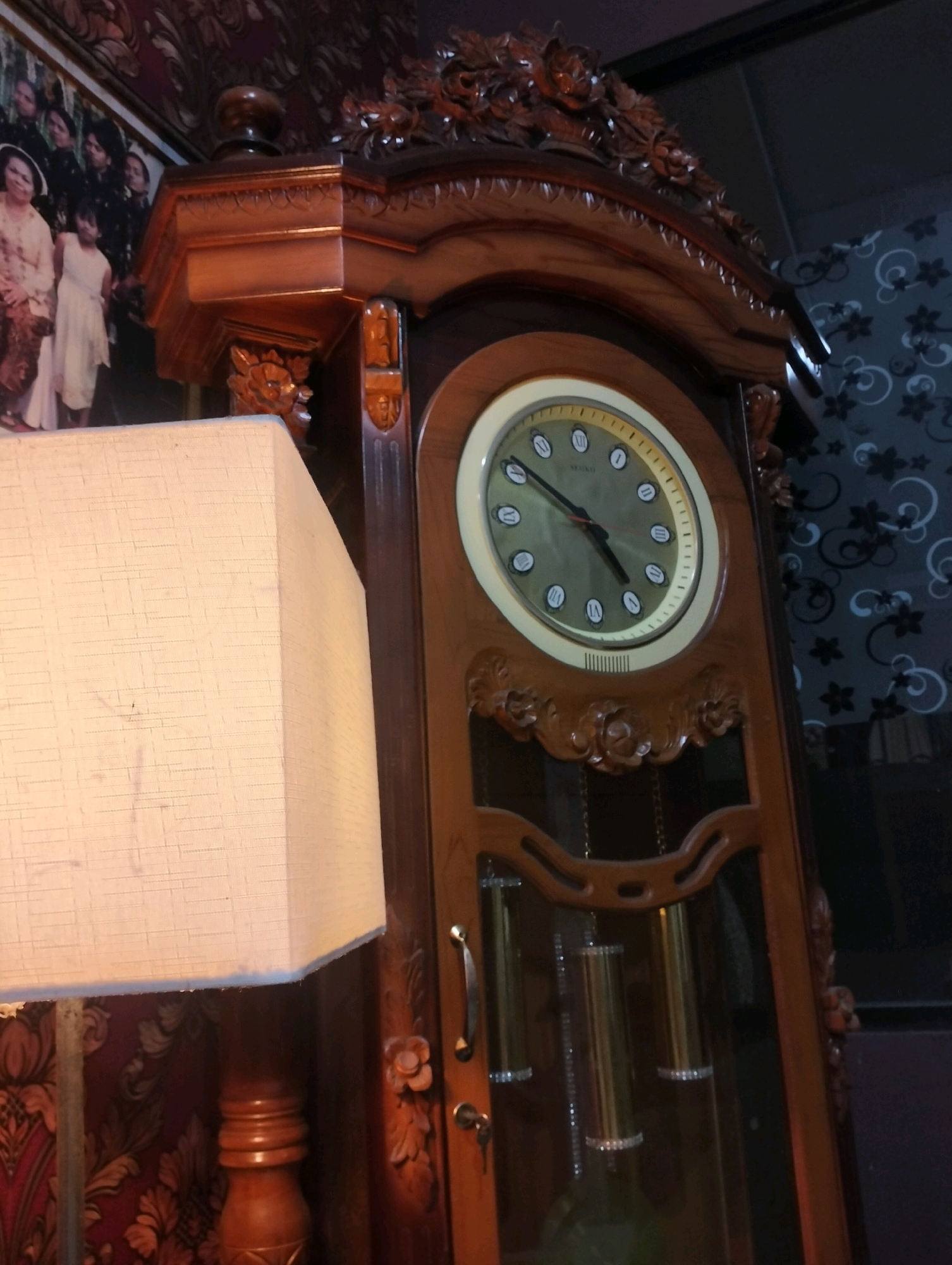 In an indoor scene, a grandfather clock with a wooden casing and a round face displaying Roman numerals is prominently featured. The clock face has black hands and a light-colored rim. Decorative carvings of flowers and foliage adorn the top and front of the clock. To the left of the clock, a tall, cream-colored lampshade with a textured surface is visible, resting on a wooden lamp base with a fluted design. In the background, a framed photograph depicts several individuals, some wearing traditional clothing. The wall behind the clock features a dark wallpaper with a repeating floral pattern. The overall impression is one of a vintage or antique setting. The location is identified as Sragen, Indonesia.