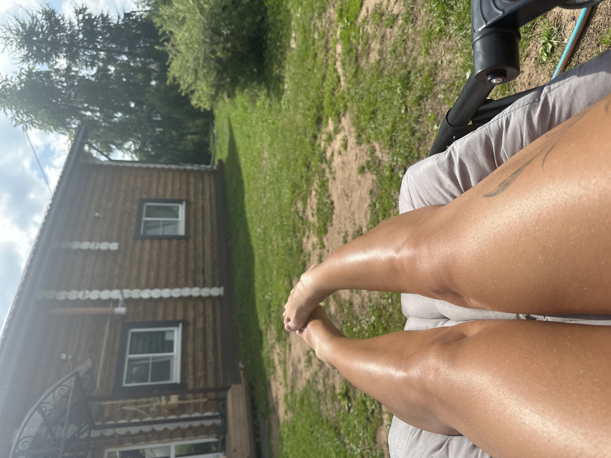 A first-person perspective shot captures tanned legs stretched out on a gray lounge chair. The legs are positioned facing upwards, with feet crossed at the ankles. The scene appears to be outdoors, with a wooden house visible in the background. The house has a gabled roof and two windows. The surrounding environment includes green grass, trees, and a cloudy sky. The location is identified as Olkhovka, Russia.