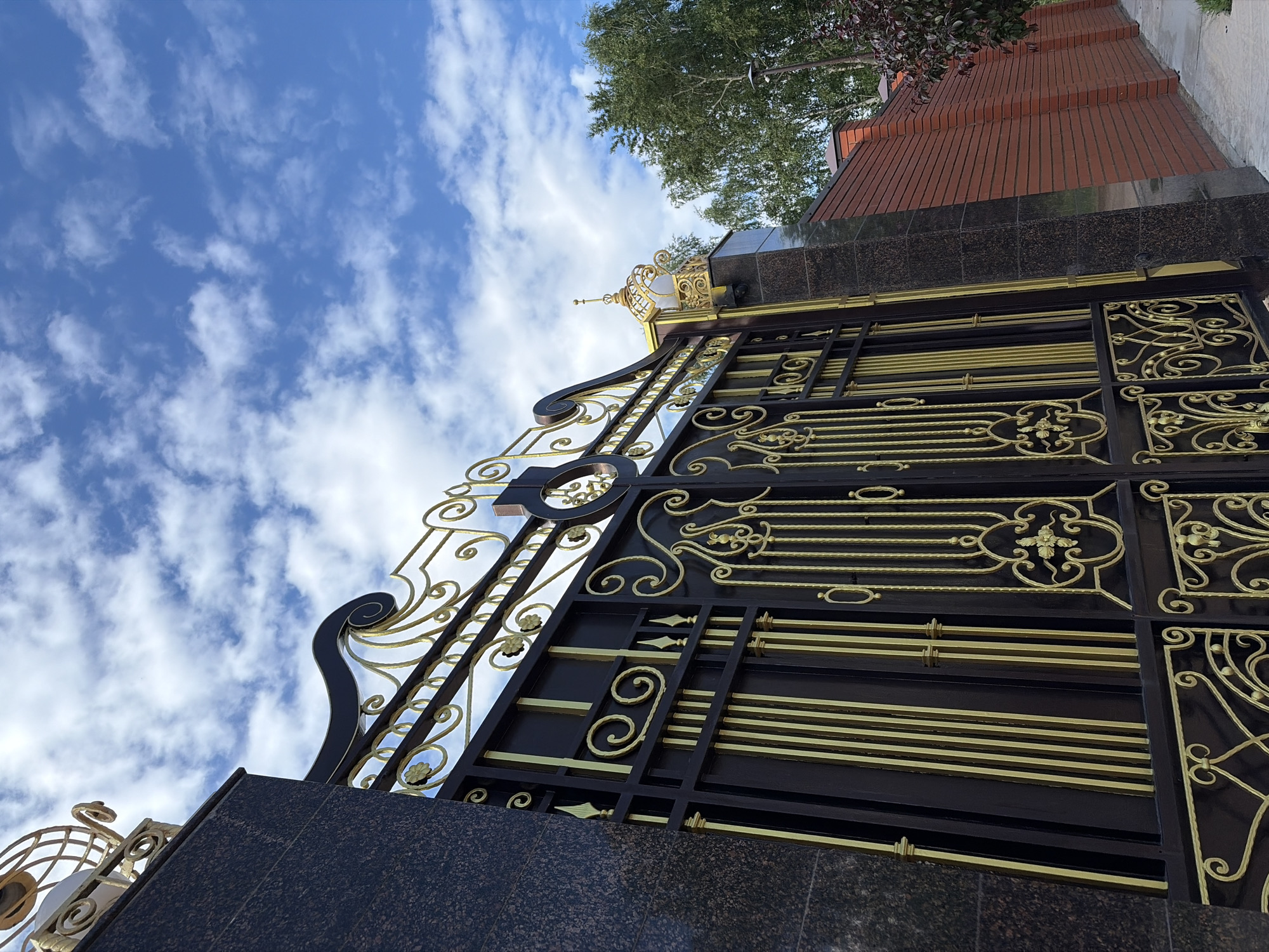 The image, captured from a low angle looking upwards, showcases a grand, ornate gate or fence against a bright blue sky. The gate is primarily dark, likely black or deep brown metal, elaborately decorated with extensive golden accents. These gold elements form intricate scrollwork, classical patterns, and stylized floral motifs, creating a striking contrast with the dark structure. The gate rests upon a sturdy base made of dark, possibly granite or marble, stone.
Above the gate, the sky is a vibrant blue, dotted with scattered, wispy white clouds, indicating a clear, sunny daytime. To the upper right, behind the gate, a structure built from reddish-brown bricks is visible, possibly a building or a high wall, partially obscured by green tree foliage. The overall setting is an outdoor architectural scene, suggesting a grand entrance or property boundary in Chaikovsky, Russia. No people or specific activities are depicted. The photograph emphasizes the scale, detailed craftsmanship, and opulent design of the gate. No visible text is present.