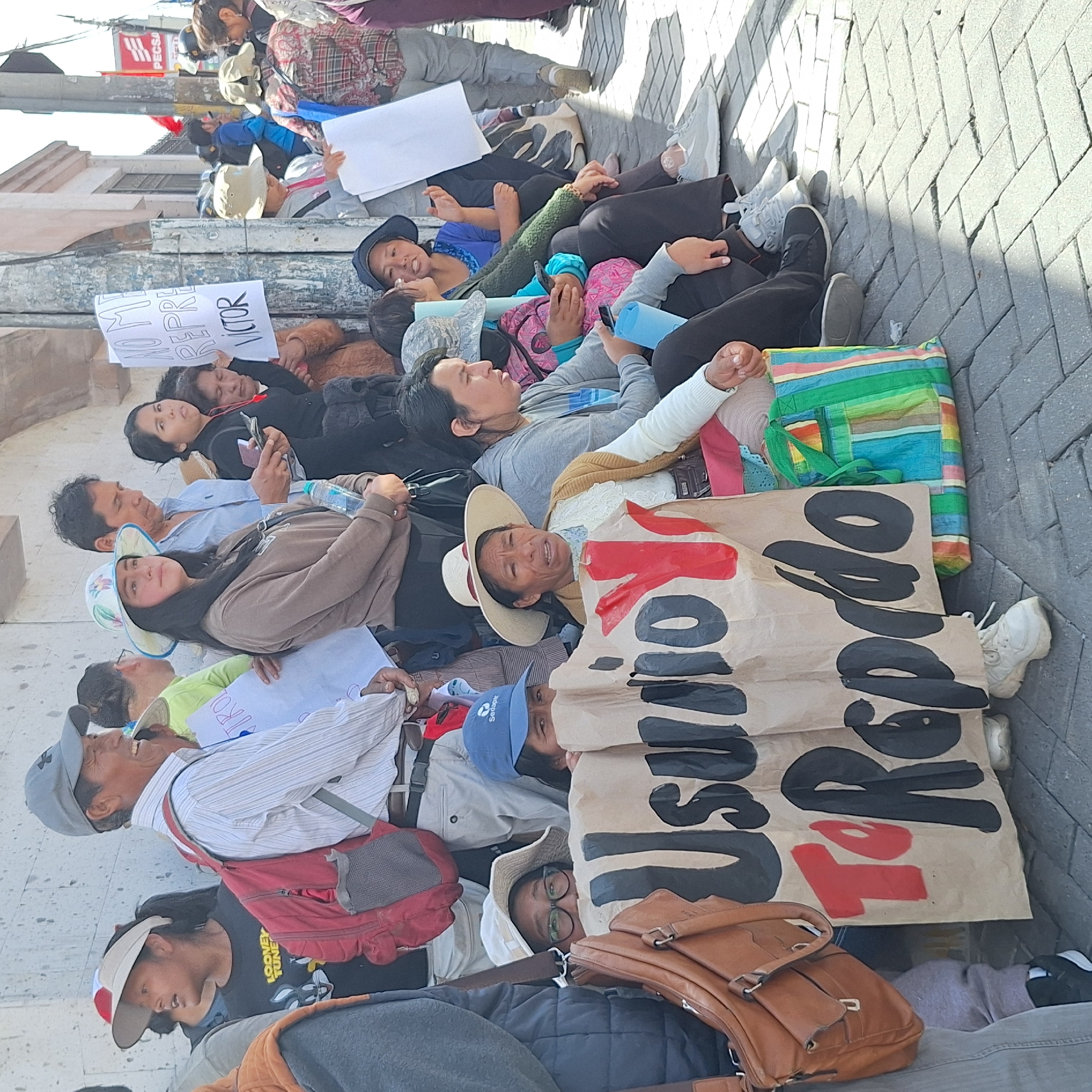 In the city of Miraflores, Peru, a group of approximately 17 individuals are lying on a gray brick sidewalk, likely engaging in a protest. The crowd is a mix of genders and ages, with participants wearing casual clothing, hats, and carrying bags. Some individuals are looking up, while others are looking at their mobile phones. Several hand-written signs are displayed, one reading "No me representa VICTOR," and another larger sign saying "INSURRECCIÓN". The protest occurs in an urban area with buildings visible in the background.