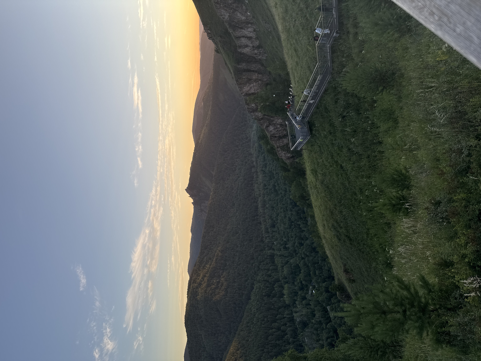 The image depicts a sunset view from a high vantage point, likely a mountain or hill, overlooking a vast, forested valley and rolling hills. The sky is a gradient of soft blues and yellows, with wispy clouds. In the foreground, a green grassy slope leads down to a wooden walkway with metal railings, where several people are seen strolling and observing the scenery. The walkway follows the contour of the hill, with stairs and landings. Further in the distance, the valley is densely covered with trees, and beyond that, layers of hills stretch towards the horizon, bathed in the warm glow of the setting sun. The overall impression is one of natural beauty and a popular tourist spot, possibly within the Stolby Nature Reserve, known for its dramatic rock formations and expansive landscapes, located near Krasnoyarsk, Russia. The scene captures a serene moment at the end of the day, with the landscape illuminated by soft, diffused light.
