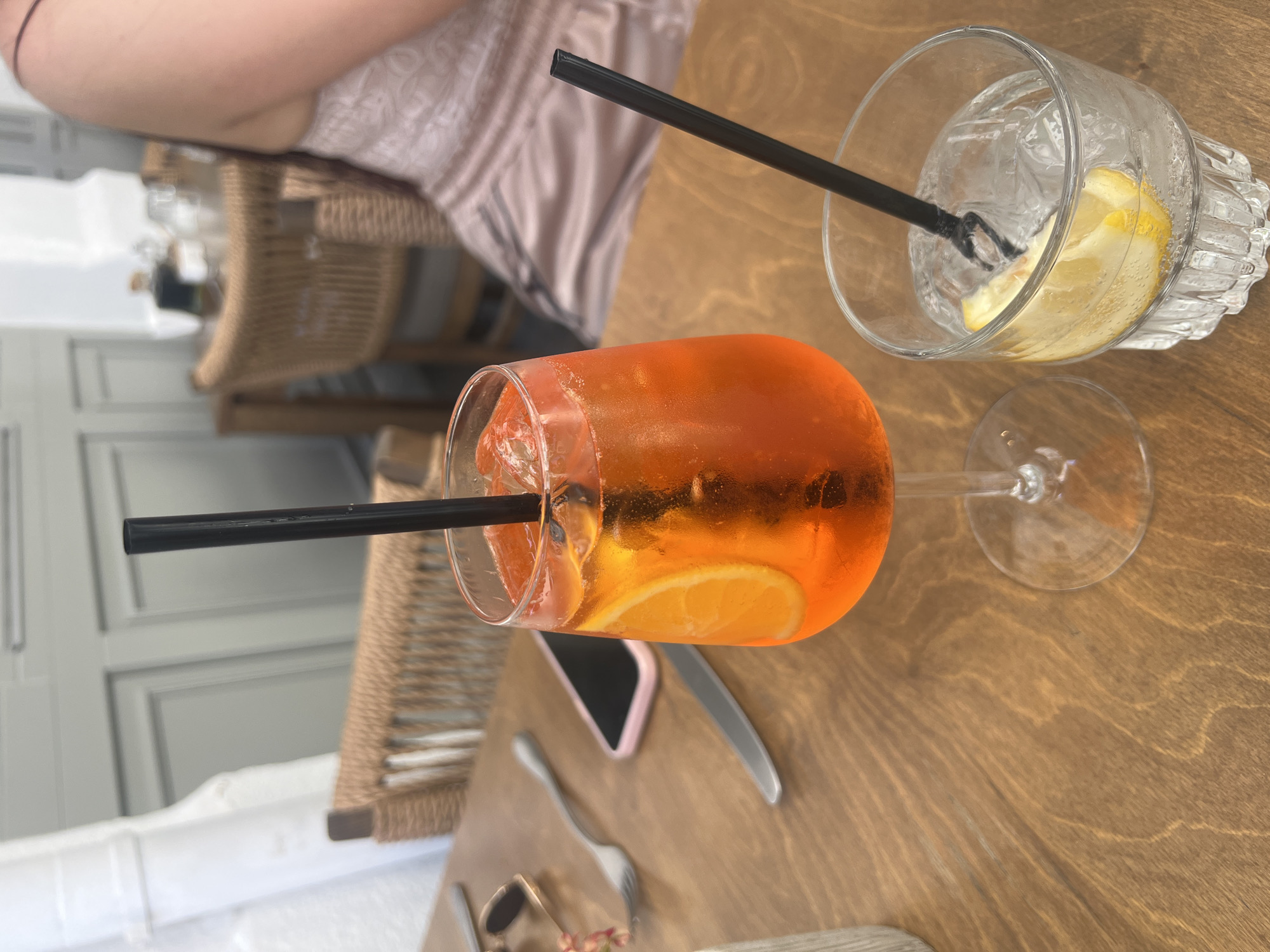 The image captures a casual tabletop scene, likely at a cafe or restaurant in Plovdiv, Bulgaria, during the daytime. Two drinks are the central subjects, positioned on a light brown wooden table with a visible grain.
On the left, an orange-colored spritz, likely an Aperol Spritz, is presented in a stemmed glass. It contains ice cubes and a slice of orange, with a black straw inserted. To its right, a clear drink, possibly water or a gin and tonic, is in a textured, faceted tumbler. This drink also contains ice and a prominent lemon wedge, along with another black straw.
A person's arm and a portion of their upper torso are visible in the upper left of the frame, wearing a light-colored, possibly lace or textured fabric top. Partially visible behind the spritz glass is a wicker chair, suggesting a comfortable seating arrangement.
Other objects on the table include a pink-cased smartphone, a silver knife, a fork, and a pair of sunglasses. The background is blurred, showing light grey or white paneled walls or structures, indicating an indoor or covered outdoor setting. The bright lighting suggests it is daytime. No discernible text is visible anywhere in the image. The overall impression is one of relaxation and leisure, with individuals enjoying beverages.