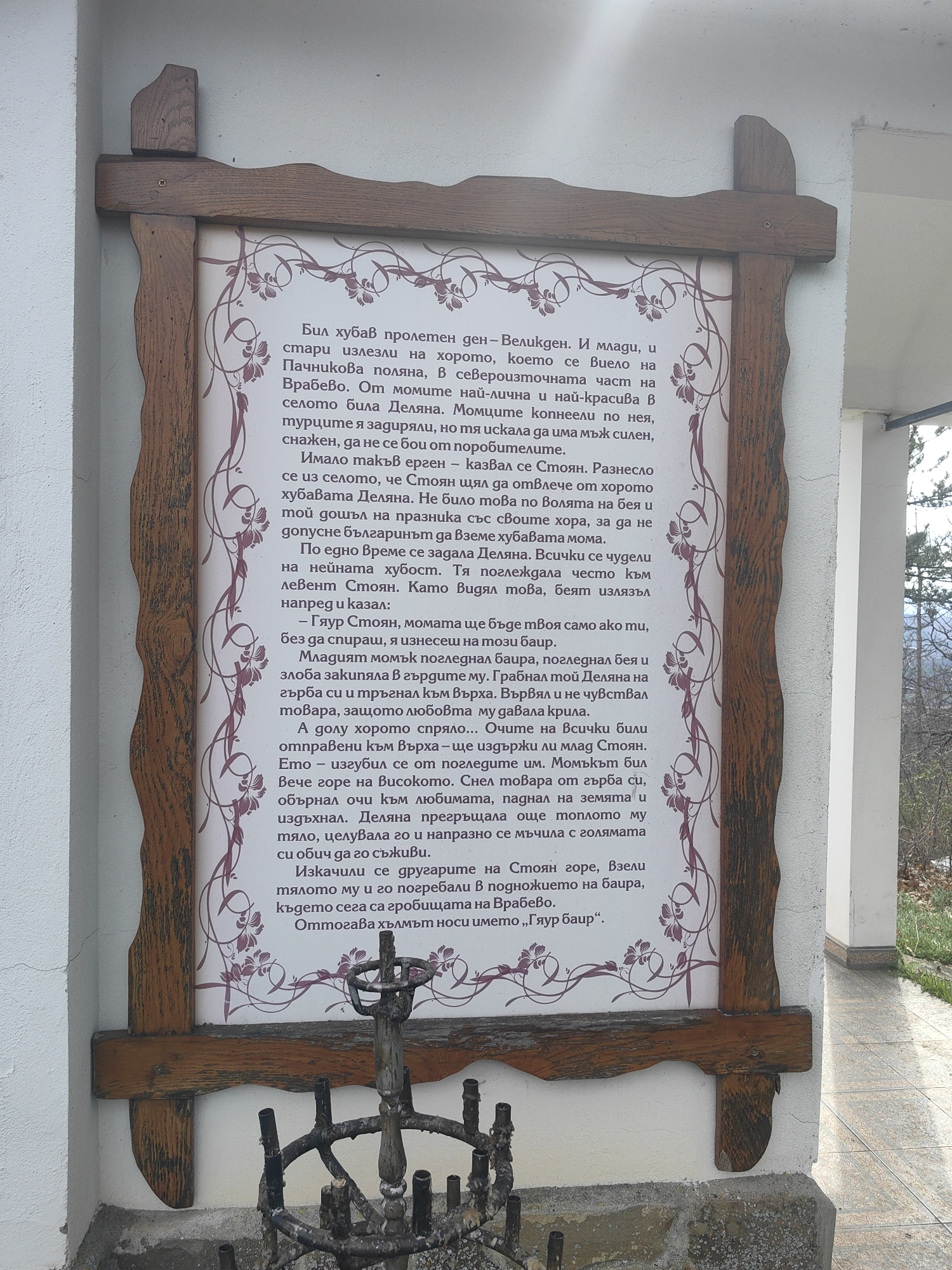 The image shows a framed text in Bulgarian, likely a historical account or legend, mounted on a wall in Gumoshtnik, Bulgaria. The text frame is ornate with wooden carvings. The text within the frame is adorned with floral designs. Below the frame is a metal structure, possibly a memorial or a religious artifact with candle holders. The text itself tells a story that describes a folk tale or historical event related to a location. In the background, trees and foliage give context to the location.