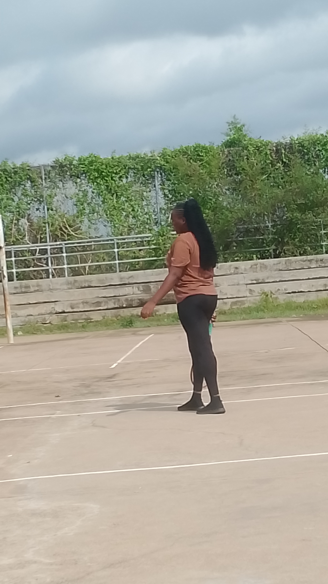 An outdoor scene in Lokoja, Nigeria, depicts a female individual standing on a light-colored concrete sports court marked with white lines. On the left, a white vertical pole, characteristic of a net support, is visible. The individual, positioned in the right half of the frame, has dark skin and long, dark braided hair tied into a high ponytail. She is dressed in a short-sleeved, rust-brown t-shirt, black leggings, and dark high-top athletic footwear. Her body is oriented in a three-quarter profile, facing towards the right side of the frame, with her head turned to look in that direction. Her left arm is slightly bent at the elbow, while her right arm is mostly obscured. In the mid-ground, behind the court, a concrete structure resembling tiered seating or a low retaining wall is present. Above this, a metal fence is heavily covered with dense green foliage. The sky overhead is uniformly overcast with grey clouds.