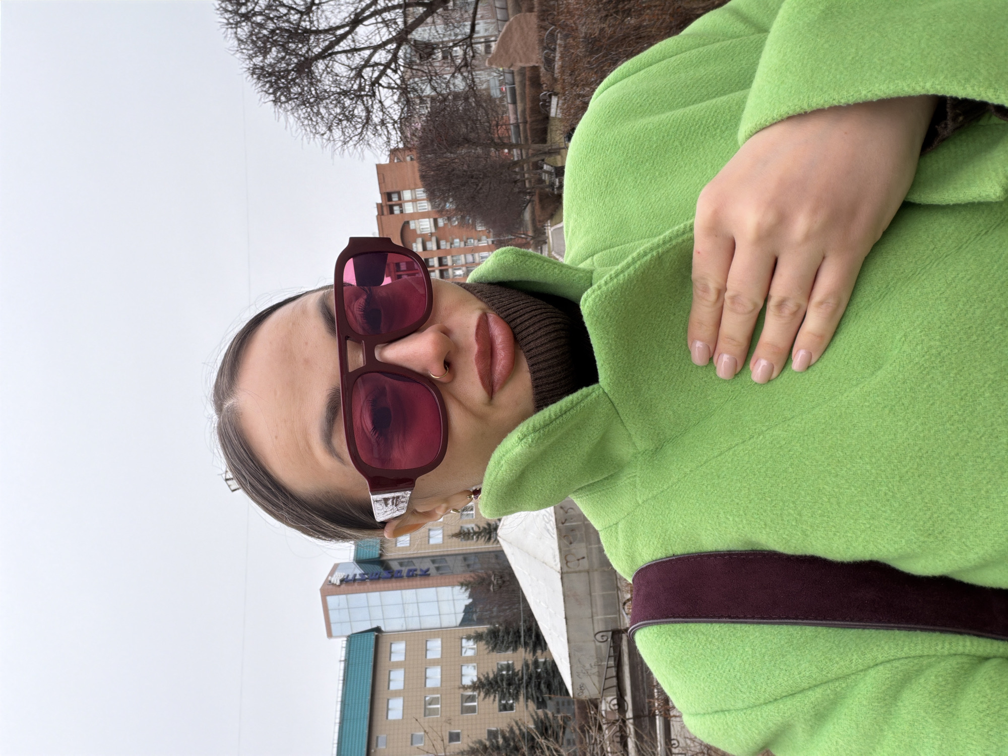 A woman is photographed outdoors in Krasnoyarsk, Russia. She wears oversized, burgundy-framed sunglasses and a bright green wool coat with a dark brown or burgundy strap across the body. Underneath, she wears a brown turtleneck sweater. She has fair skin, with a nose ring and gold earrings. Her hand rests on her coat collar.
In the background, there are buildings, trees, and a cloudy sky. One of the buildings has a blue-green and white facade. Bare tree branches are visible against the skyline. The scene suggests it is likely a cold or overcast day.