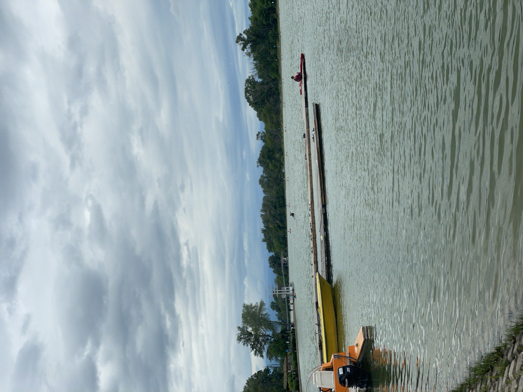 The image shows a lakeside view in Plovdiv, Bulgaria. A body of water stretches across the frame, reflecting a cloudy sky. A wooden dock extends into the water, with two boats moored nearby. A person is visible in a kayak further down the dock. Trees line the opposite bank of the lake, and a lifeguard stand is seen on the shoreline. An outboard motor is visible on one of the boats.