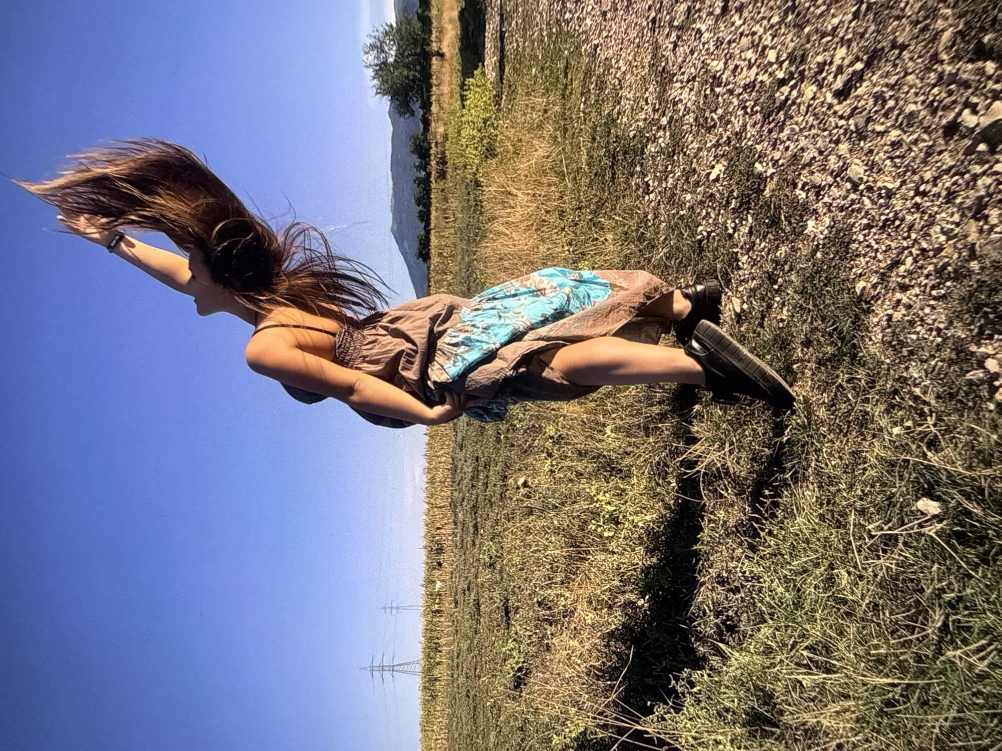 A woman is standing in a field on the outskirts of Sofia, Bulgaria. She is wearing a brown dress with blue accents and black shoes. Her long brown hair is thrown forward as she reaches towards the sky. The field is overgrown with dry grass and some patches of green. In the background are distant hills and power lines, suggesting the rural-urban fringe of the city. The angle is tilted, creating a dynamic perspective.