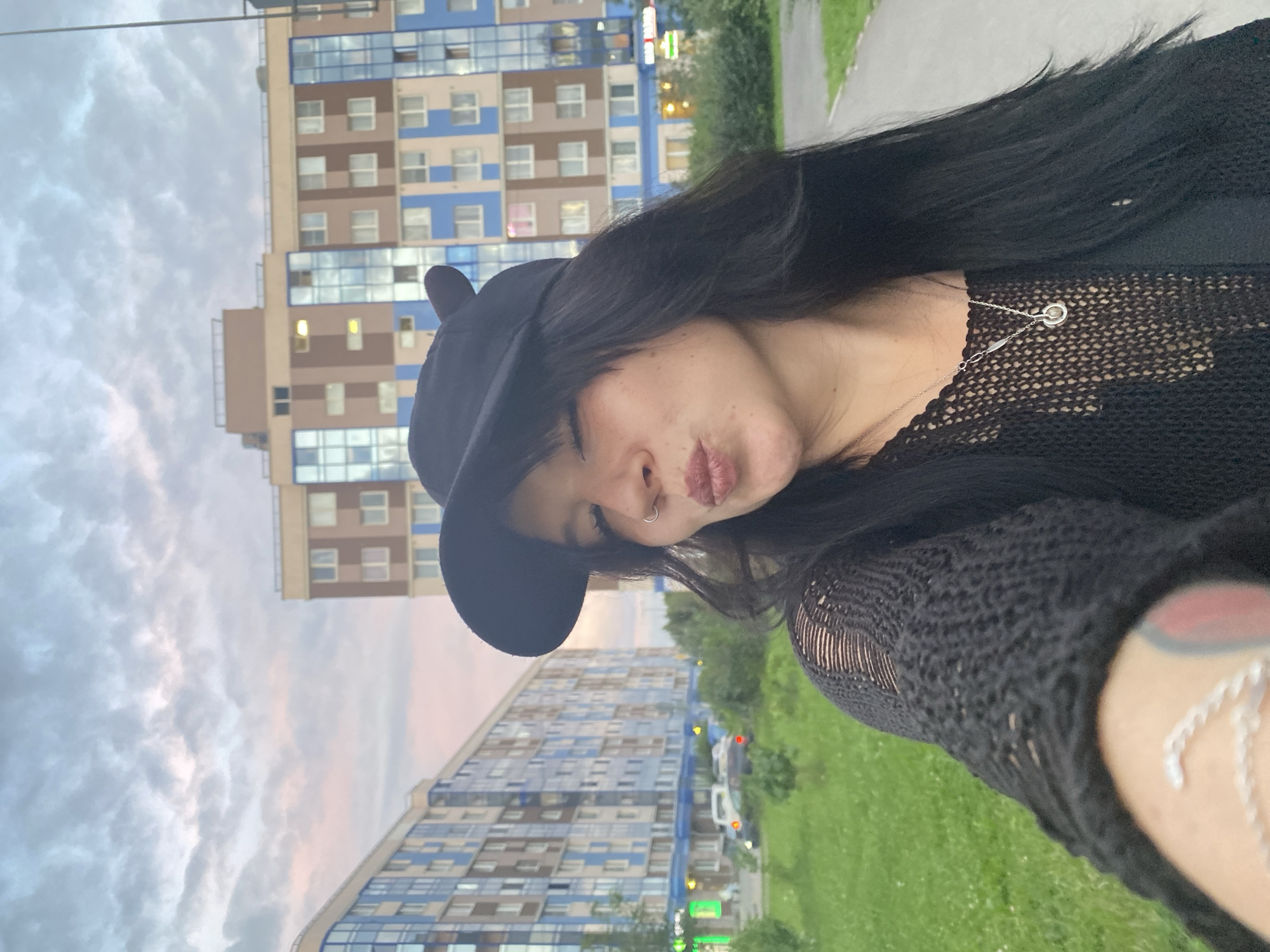 The image is a selfie taken by a young woman with long, dark hair, wearing a black knit cardigan over a dark top, a black baseball cap with ear details, and a silver necklace. She has a nose ring and is making a kissing face towards the camera. She appears to be outdoors in front of apartment buildings in Yanino-1, Russia. The buildings are multi-story residential complexes with a mix of beige, brown, and blue facades, and many windows. There is a grassy area with trees and bushes in the foreground, and a paved pathway to the right. The sky is cloudy with hints of pink and orange, suggesting it might be near sunset. The overall impression is a casual, urban setting.