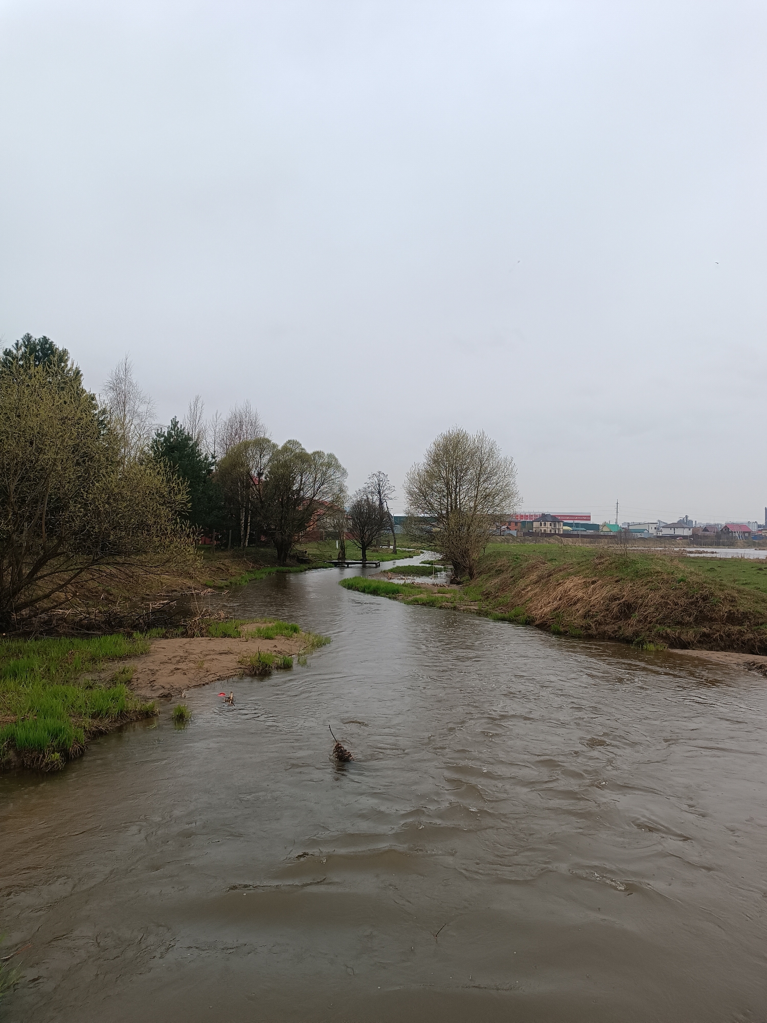 The scene depicts a flowing river in Motyakovo, Russia, under an overcast sky. The riverbanks are lined with grass, sand, and sparse trees with budding leaves, suggesting early spring. A small footbridge spans the river in the mid-ground. In the background, buildings of the village or town are visible. Debris, including a piece of litter and a clump of sticks, is present in the water.