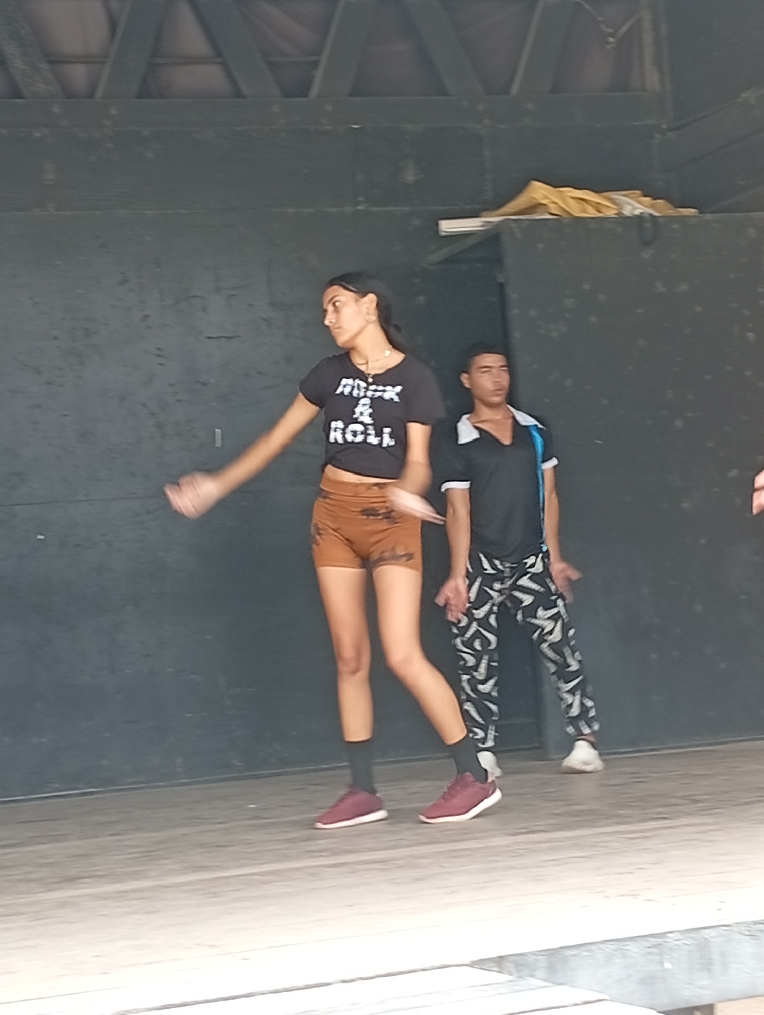 The image shows two people standing on a wooden stage in Playa El Agua, Venezuela. A young woman is on the left, wearing a black cropped t-shirt with "ROCK & ROLL" printed on it, brown shorts, black socks, and maroon sneakers. Her hair is dark, and she has a necklace. She appears to be dancing, with her left arm extended. A young man is standing behind and to the right of her, wearing a black collared short-sleeve shirt with a light blue stripe, camouflage-patterned pants, and white sneakers. He also appears to be dancing or posing. The background is a dark, slightly weathered wall. Above and behind them, there is what appears to be a roof structure with wooden beams. A light-colored tarp is visible on top of the structure. The stage is constructed of light-colored wood.