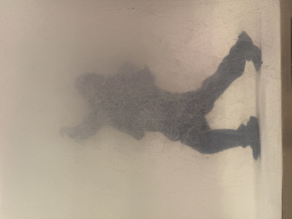 The image displays a static art installation viewed through a translucent, heavily scratched protective panel. A single human figure is represented as a dark, dense, fibrous silhouette, appearing fuzzy in texture. The figure is captured in a dynamic pose, leaning forward with the left leg extended in front and the right arm raised and bent, suggesting an action of throwing or forceful forward motion. The head is slightly angled down and to the right. The background behind the figure and panel is a uniform, light, off-white hue, indicating an illuminated interior space, with the figure appearing to stand on a similarly light-colored ground plane. This artwork is part of "The Swarm" installation by Miroslav Yotov, situated in the pedestrian underpass connecting the Presidency and the Council of Ministers building, located near the Serdica metro station complex in the city center of Sofia, Bulgaria.