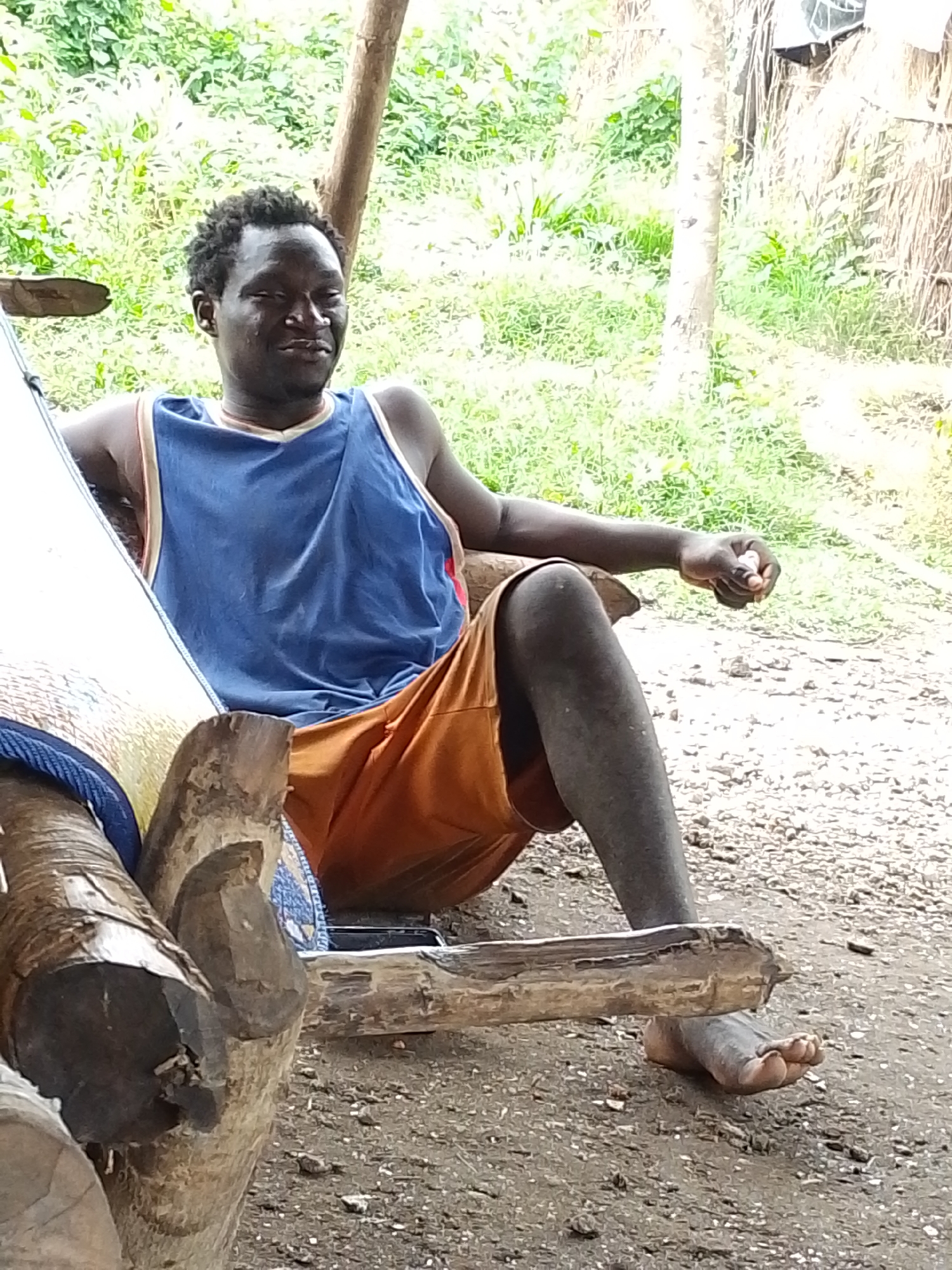 A man sits in a wooden chair outdoors in Otulu, Nigeria. He is wearing a blue tank top and orange shorts. The man is holding something in his right hand and appears to be looking down at it. The background is a blur of green foliage and trees. The ground is dirt and gravel.