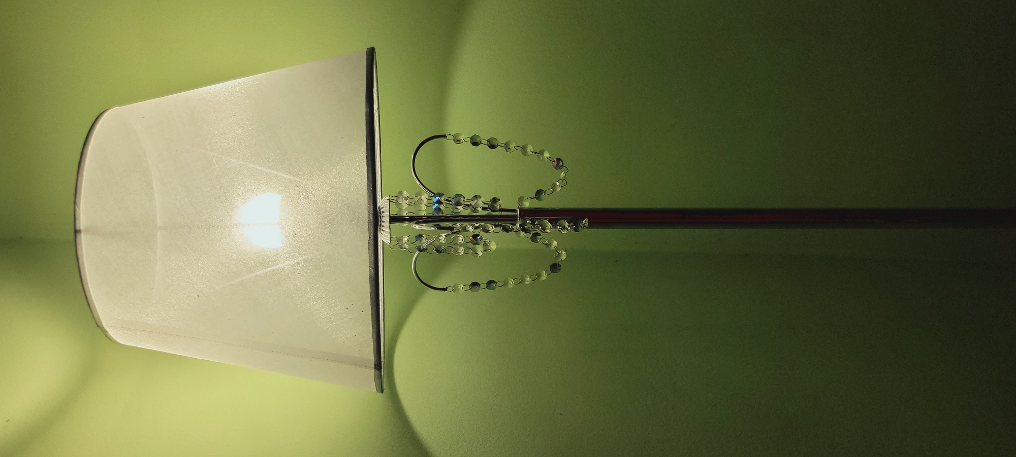 An interior shot focuses on the upper portion of a floor lamp against a lime green wall. The lampshade is a translucent white, a warm light bulb is visible within, casting a glow. Attached to the lampshade's fixture is a decorative chain of clear and faceted beads, some with hints of blue and purple. The lamp's slender metallic pole extends downwards out of frame. The wall shows subtle texture and shadows cast by the lamp. The scene is indoors, with no indication of a specific city or location beyond "Unknown, Unknown."