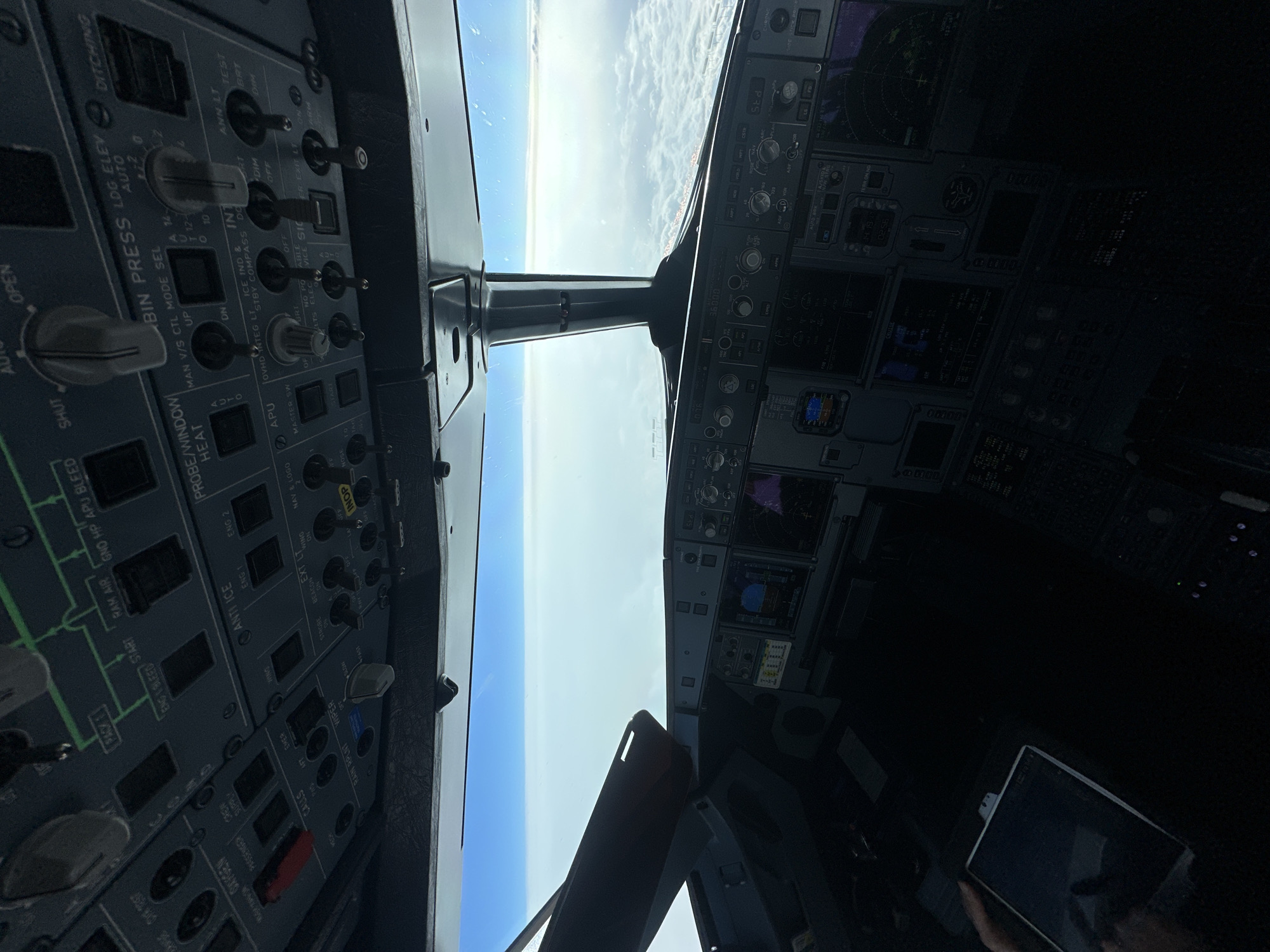 The image shows the cockpit of an airplane in flight, likely an Airbus based on the panel layout. The view out the front window shows a sky with blue sky and clouds. The left panel displays various controls including engine start, anti-ice, probe heat, and bin pressure settings. The right panel is darker but shows some flight instruments and displays. An electronic tablet is visible in the lower right, likely being held by a person whose hand is partially visible. The image does not contain any indications that would suggest the location is Sochi, Russia.