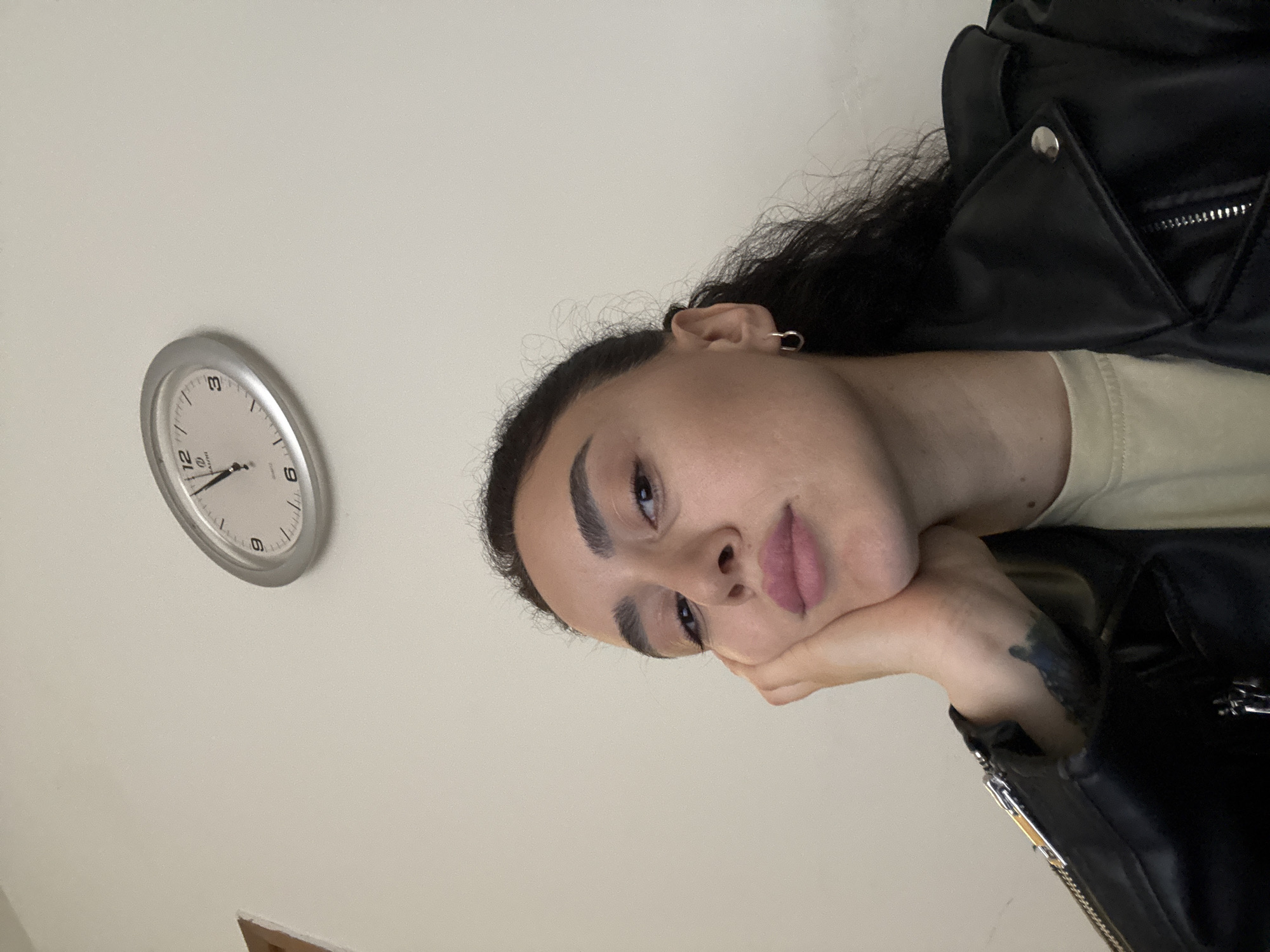 A person is seen in a low-angle portrait against a light-beige wall. The individual's face is the central focus, with the subject resting their cheek on a hand that bears a dark tattoo. They have dark hair, well-defined eyebrows, and lips with neutral-toned lipstick. The person is wearing a black leather jacket over a light-colored shirt. A silver-framed clock is visible on the wall, with the hands indicating the time just past 11:00. There is no discernible information in the image to identify the location as Tbilisi, Georgia or any specific urban environment.