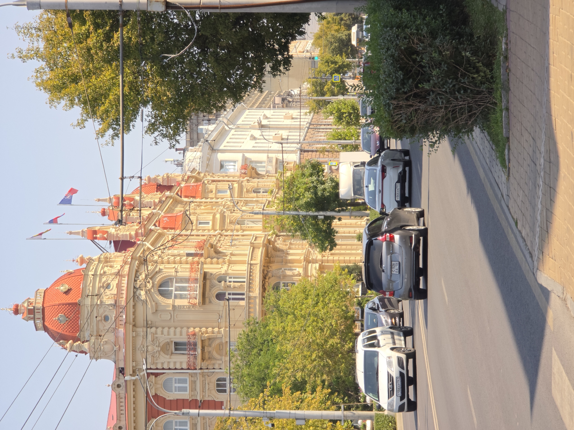 The image shows a street scene in Rostov-on-Don, Russia, on a sunny day. The foreground features a paved road with several cars driving on it, including a white van, a gray hatchback, and a white sedan. A pedestrian crossing is visible on the left side of the road. The middle ground displays a row of trees with lush green foliage lining the street. In the background, an ornate, light-colored building with red-tiled domes and red accents dominates the scene. This building is likely a historical landmark or a significant architectural structure in the city. Several flags, including what appear to be Russian flags, are visible atop the building. Power lines crisscross the sky above the street and building. The overall impression is a bustling urban environment with a mix of modern traffic and historical architecture.
