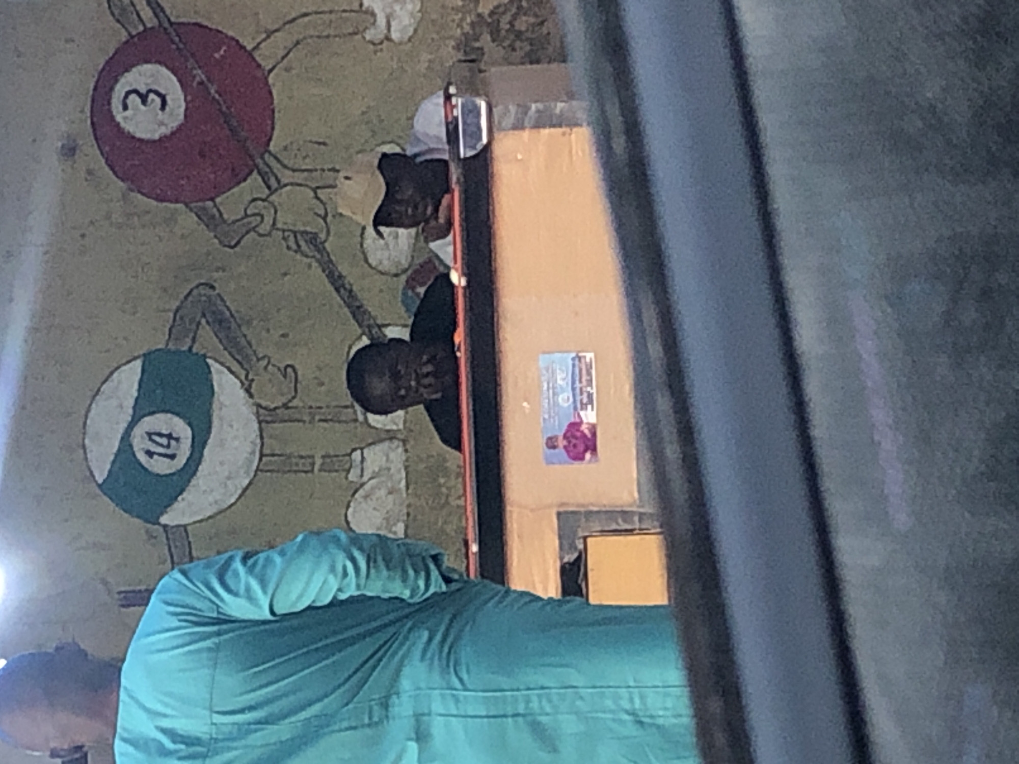 In Unknown, Unknown, a street-level view captures three individuals interacting around a game table against a wall mural. The background wall, light-colored, features two large, cartoonish depictions of anthropomorphic pool balls: one striped white and green with the number "14", and another solid red with the number "3" holding a pool cue. In the foreground, a bald individual, seen from the back, wears a loose-fitting, long-sleeved teal garment. They stand facing a light-brown wooden-based game table. Two dark-skinned individuals are positioned at the table; one on the left, wearing a dark shirt, has hands near their face, while the other on the right, wearing a white t-shirt and a light-colored baseball cap, leans on the table with a pool cue visible to their right. A small poster with a person in a red shirt is affixed to the side of the table base. The scene is illuminated by an overhead light source, creating a bright spot in the upper left.