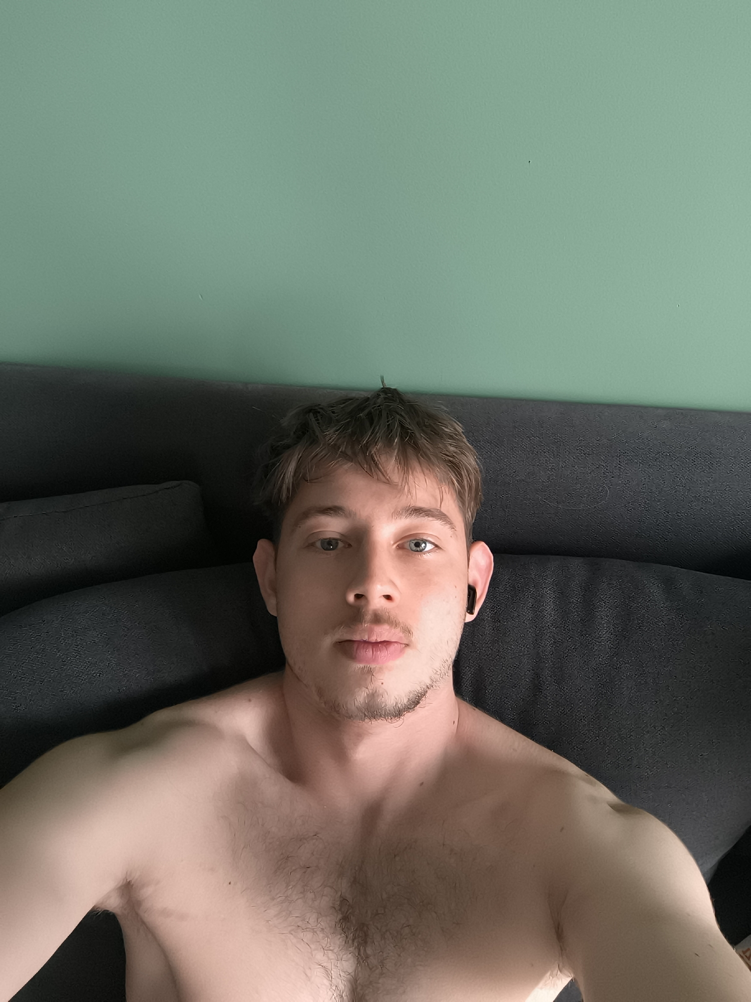 A topless man with fair skin, blue eyes, and short brown hair lies on a dark grey sofa against a pale green wall. He has stubble on his face and is wearing a black earbud in his right ear.
It is not possible to determine the exact location of this scene in Sofia, Bulgaria, based on the image alone.