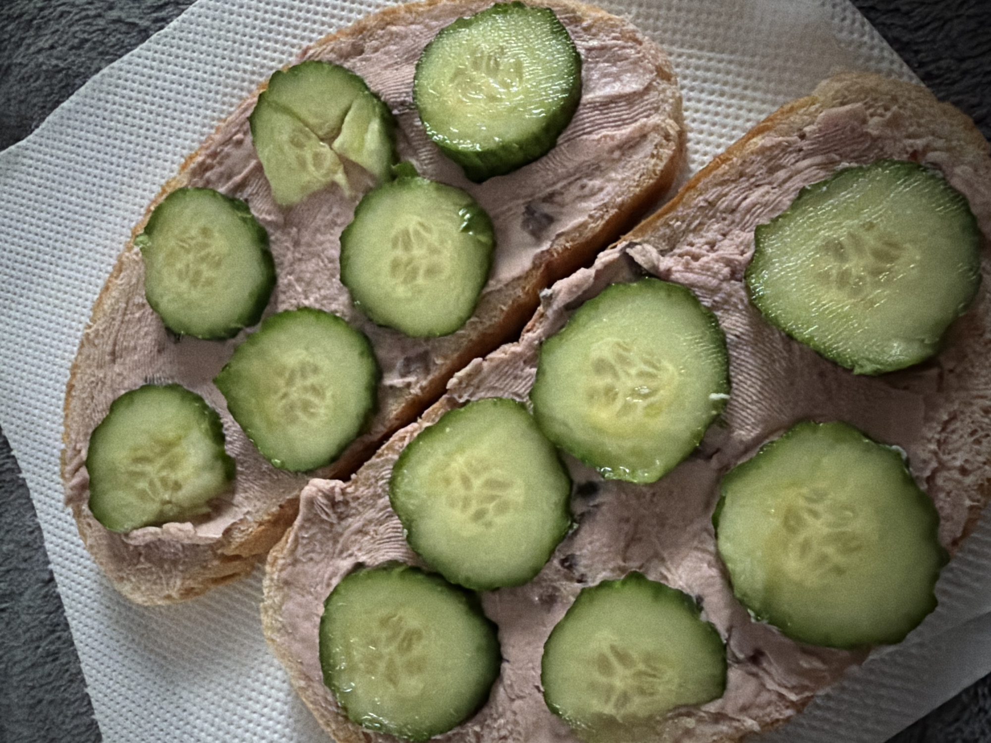 Two slices of bread, spread with a pinkish-brown pate, are topped with sliced cucumbers and arranged on a white paper napkin. The napkin rests on a gray surface. The image does not provide enough information to determine the location in the city of Sofia, Bulgaria, or any information about people, subjects, actions, interactions, or specific environmental details beyond what is described.