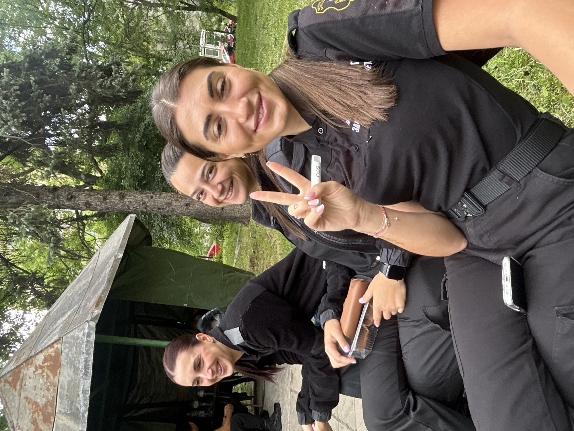 Three young women in black uniforms are posing for a selfie. The woman in the foreground is holding up a peace sign and smiling. She has a smartwatch on her left wrist and a phone in her right pocket. The other two women are smiling in the background. One is holding a package of what appears to be cookies. The women are standing outside a makeshift tent-like structure. The scene appears to be in a park, with trees and grass visible in the background. The location is likely Sofia, Bulgaria.