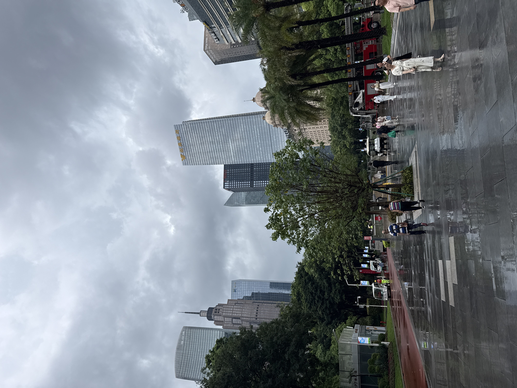 This is a full-frame, eye-level, outdoor, medium shot of a street in Tianhe District, China, on a cloudy day. The wet, dark, grey, stone pavement reflects the surrounding environment, indicating recent rain. In the foreground, two people are walking away from the camera on the right side of the frame. The woman on the right wears a light-colored, floral dress and sandals, while the person to her left wears a white top and a black skirt. Further back, a group of people walk from left to right, including a person in a white dress, another in a white top and pants, and others in casual attire. To the left, a red path is visible, bordered by a low wall. Several small, white electric vehicles are parked along the left side of the path. A red fire truck is parked on the right side of the frame, behind some palm trees. In the background, a dense cluster of modern skyscrapers dominates the skyline under a heavy, grey, cloud-covered sky. Some of the buildings have Chinese characters on them. Trees line the street, with lush green foliage. The overall atmosphere is urban and slightly damp.