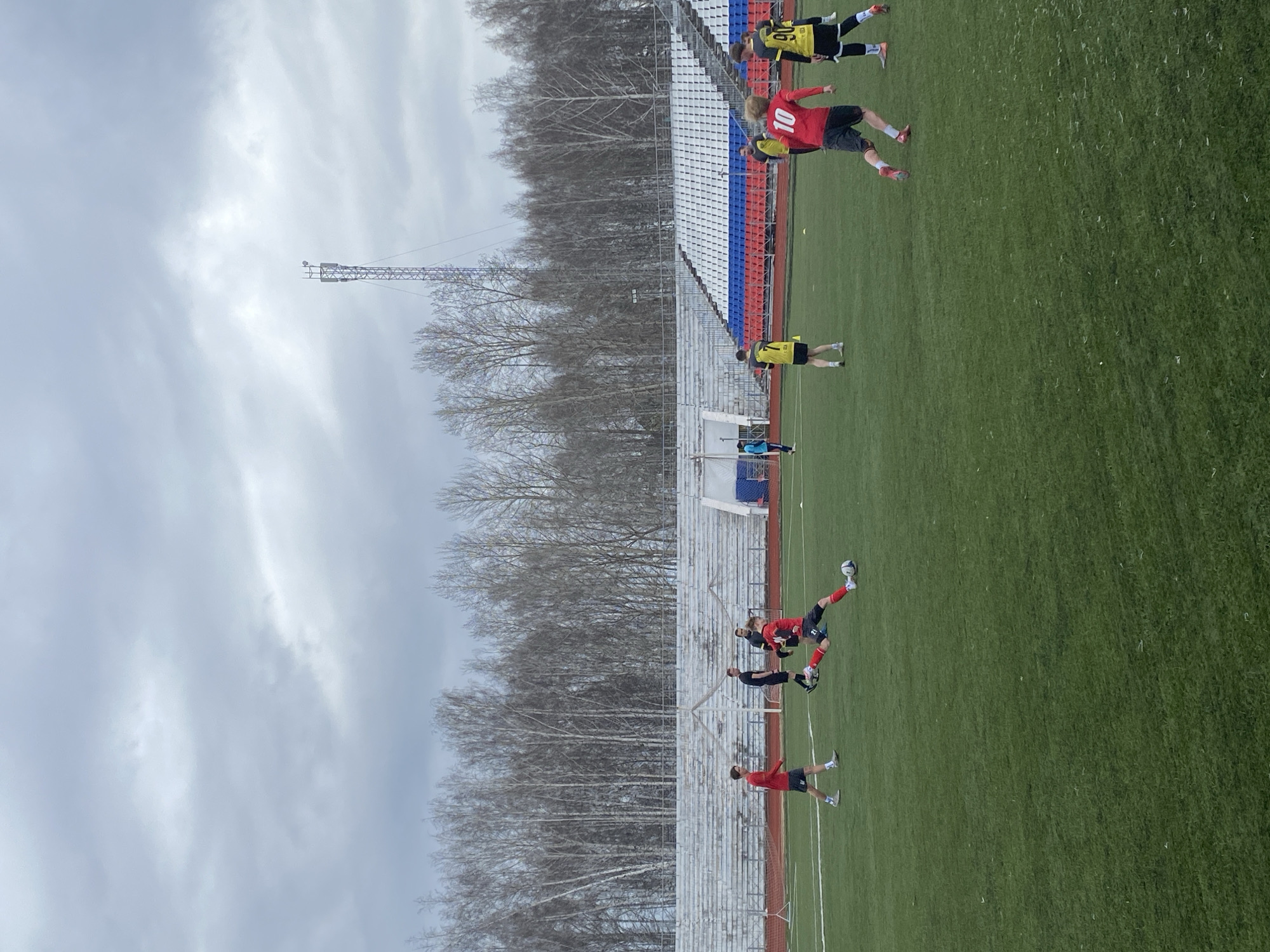 The image depicts a soccer match in Chaikovsky, Russia. Several players in red and yellow uniforms are on a green field. A soccer ball is in the air. Stadium seating with red, white, and blue sections is visible behind the field. A bare forest lines the background. The sky is overcast. A tall, metal structure is visible near the forest.