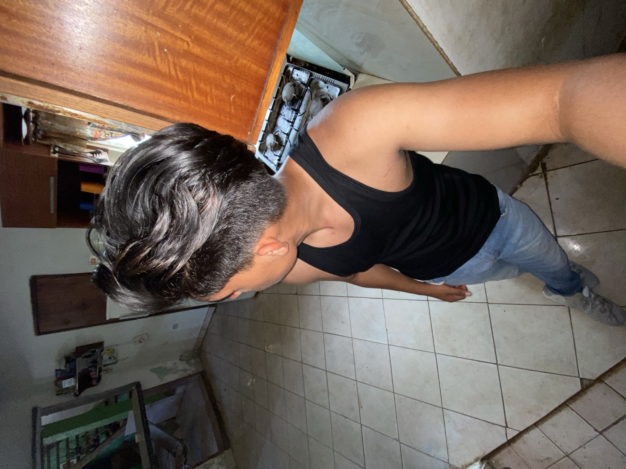 The image depicts a man standing in a kitchen area, likely located in a residential space. The man is wearing a black tank top and blue jeans. He appears to be holding a camera or phone to take a selfie. The kitchen area has tiled floors and wooden cabinets. A stove is visible next to the cabinet. The kitchen environment is worn, and the lighting is dim. There's no indication that this image is specifically from Juan Griego, Venezuela, so it is impossible to definitively confirm its location based on the visual data.