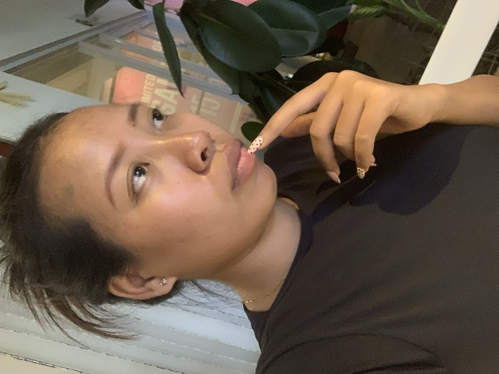 A young woman is shown in a close-up, profile view, looking off to the right. She has dark hair pulled back, and is wearing a dark t-shirt and a delicate gold necklace. Her left hand is raised to her chin, with her index finger touching her lips. Her fingernails are painted with a light pink base and black dots. In the background, a large green plant is visible, along with a window with a sign that appears to read "LIMITED SALE". The lighting suggests it is evening or indoors. The image does not contain enough information to determine its location within Denpasar, Indonesia.