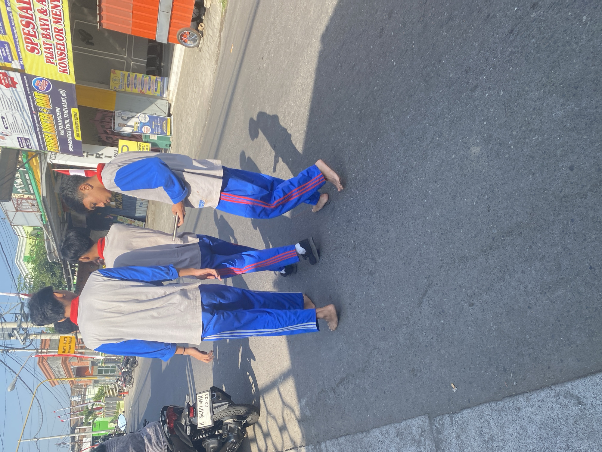 The image shows three young males walking on a paved street in Kediri City, Indonesia. The individual in the foreground is wearing a gray long-sleeved shirt with blue sleeves and blue athletic pants with white stripes. He is barefoot and holding a smartphone. To his right, another male is walking barefoot in similar attire, wearing blue pants with red stripes and a gray shirt. A third male, partially visible, is also wearing blue pants with red stripes. In the background, there are buildings with signs advertising various services, including medical clinics and health services. A motorcycle is parked to the left of the frame, and a red kiosk with a wheel is visible on the right side of the street. The scene appears to be outdoors during daylight, with shadows cast on the road, indicating sunlight.