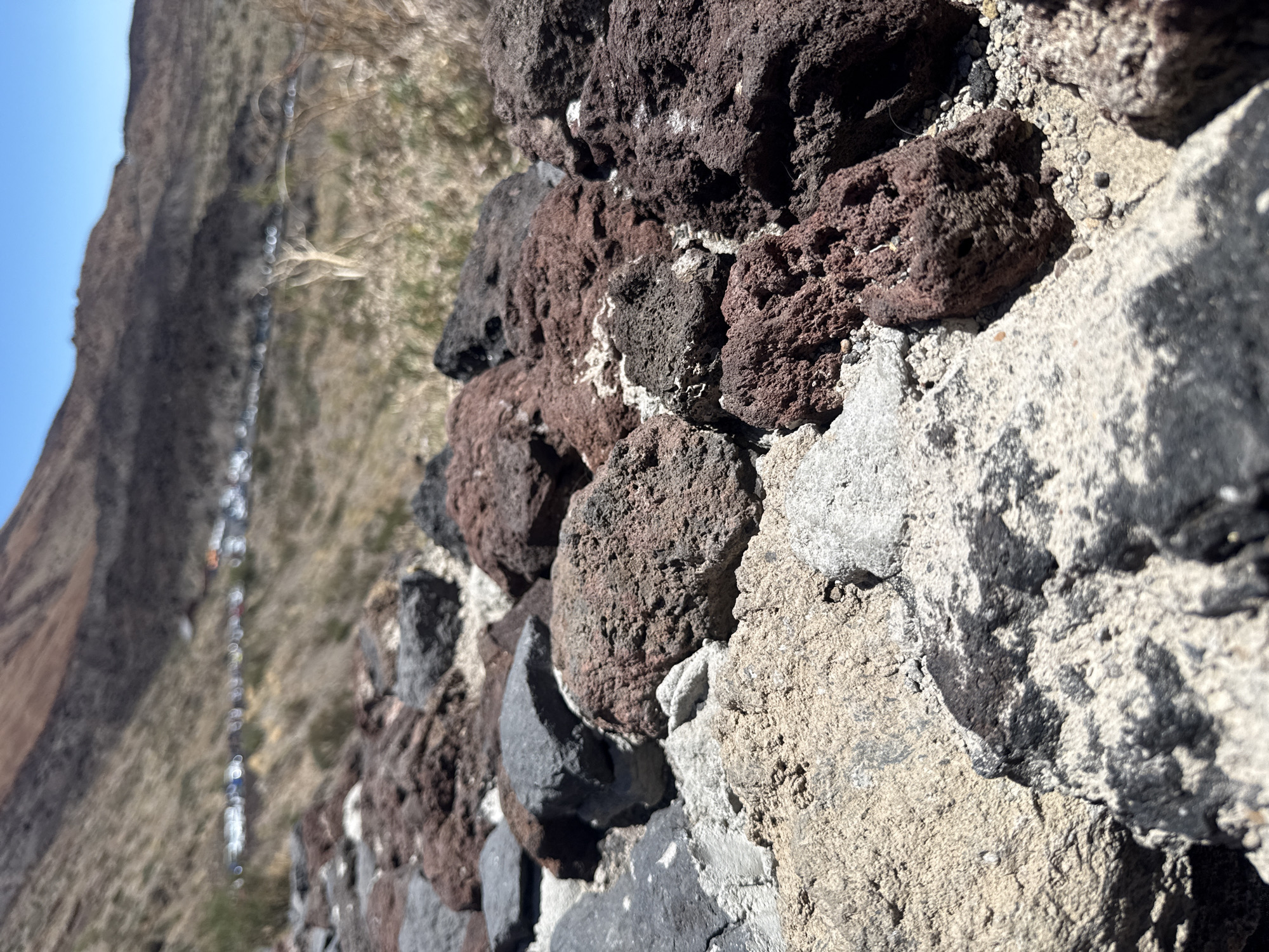 The image shows a close-up view of rough, dark volcanic rocks and lighter-colored stones arranged on the ground, possibly forming a path or a wall. In the background, a blurred landscape unfolds, featuring a barren, mountainous terrain with sparse vegetation under a clear blue sky. A line of blurred vehicles, suggesting a parking area or a road, is visible in the mid-ground. The scene is outdoors, under natural daylight. The location is identified as Teide National Park, near the city of La Orotava, Spain, a region known for its volcanic geology.
