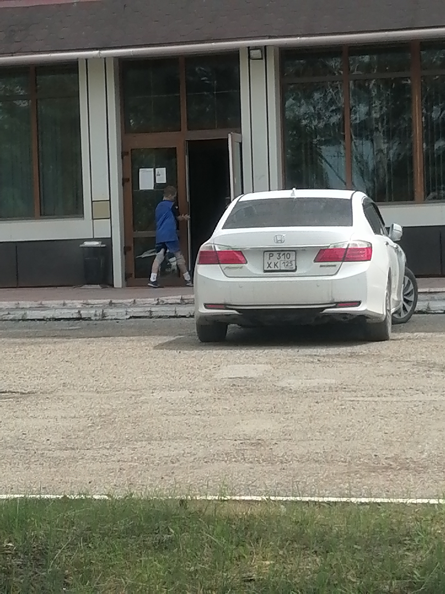 A young person wearing a blue jacket and dark shorts is exiting a building through a double glass door. The person is captured mid-stride, moving from left to right. In front of the building, a white sedan is parked, facing away from the viewer. The license plate of the car is visible and reads "P 310 XK 125". The building appears to be a commercial or public establishment with large windows and a dark roof. The ground in front of the building is a gravel surface, and there is a strip of grass at the bottom of the frame. The scene is set on a cloudy day.