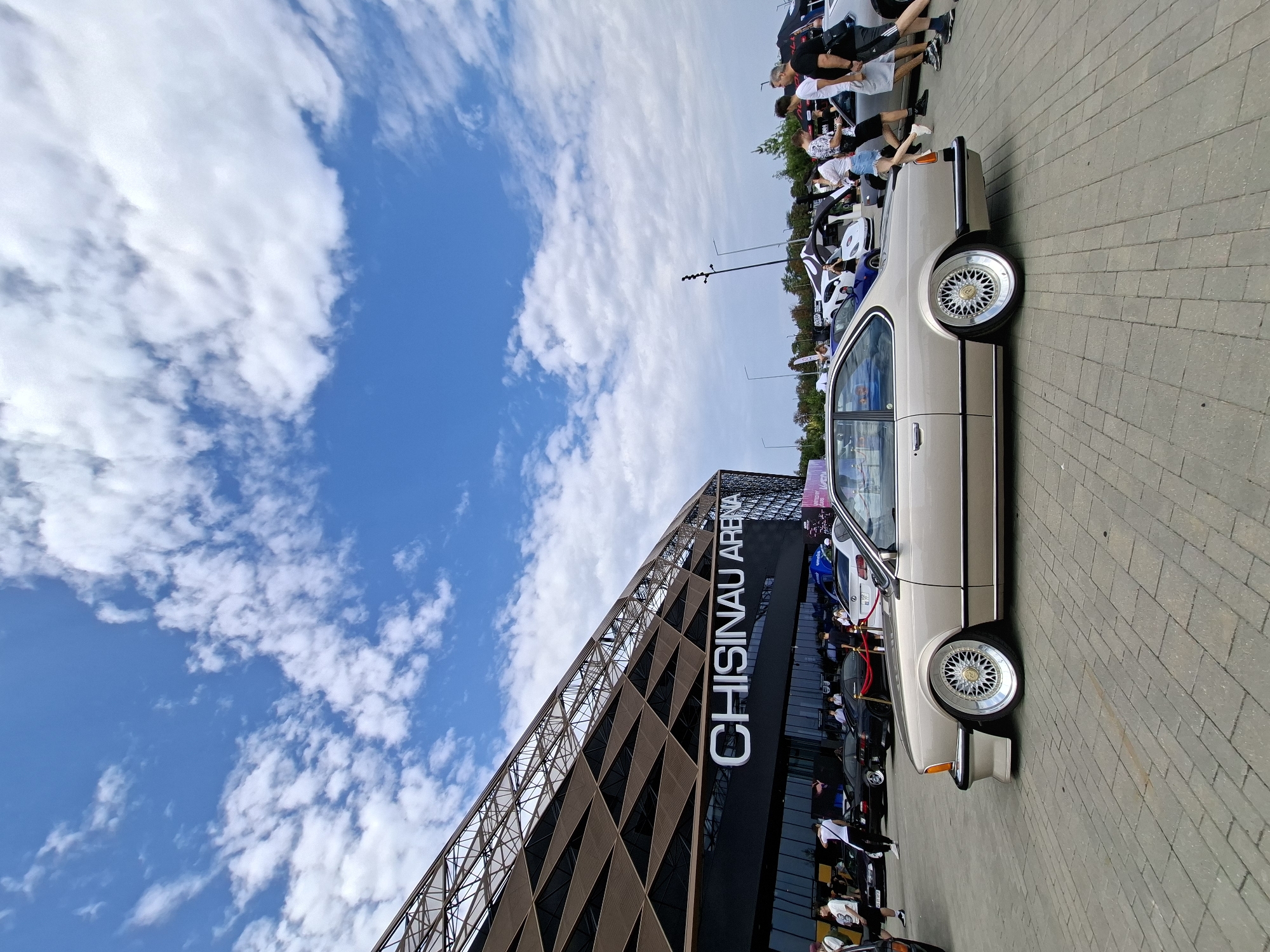A beige coupe automobile is parked on a paved surface in front of a modern building with the words "CHISINAU ARENA" in large white letters. The building has a distinctive angular roof structure with interwoven geometric patterns. Several people are visible in the background, some standing near the cars and others walking. Other vehicles are parked in the vicinity, including black sedans and a white sports car with its hood open. The sky is blue with scattered white clouds. The scene appears to be an outdoor event or car show. The location is the Chisinau Arena, located in the city of Stăuceni, Moldova.