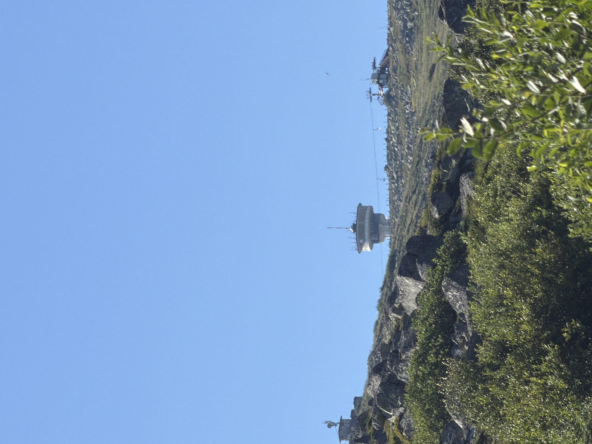 The image shows a mountainous landscape on a clear day. In the distance, on the top of a hill, there are several structures, including a round tower with balconies and a smaller building with an antenna. There also appears to be a ski lift with chairs suspended in the air. The foreground is filled with lush green bushes and large rocks. The location is identified as Bistritsa, Bulgaria. There are no people visible in the image.
