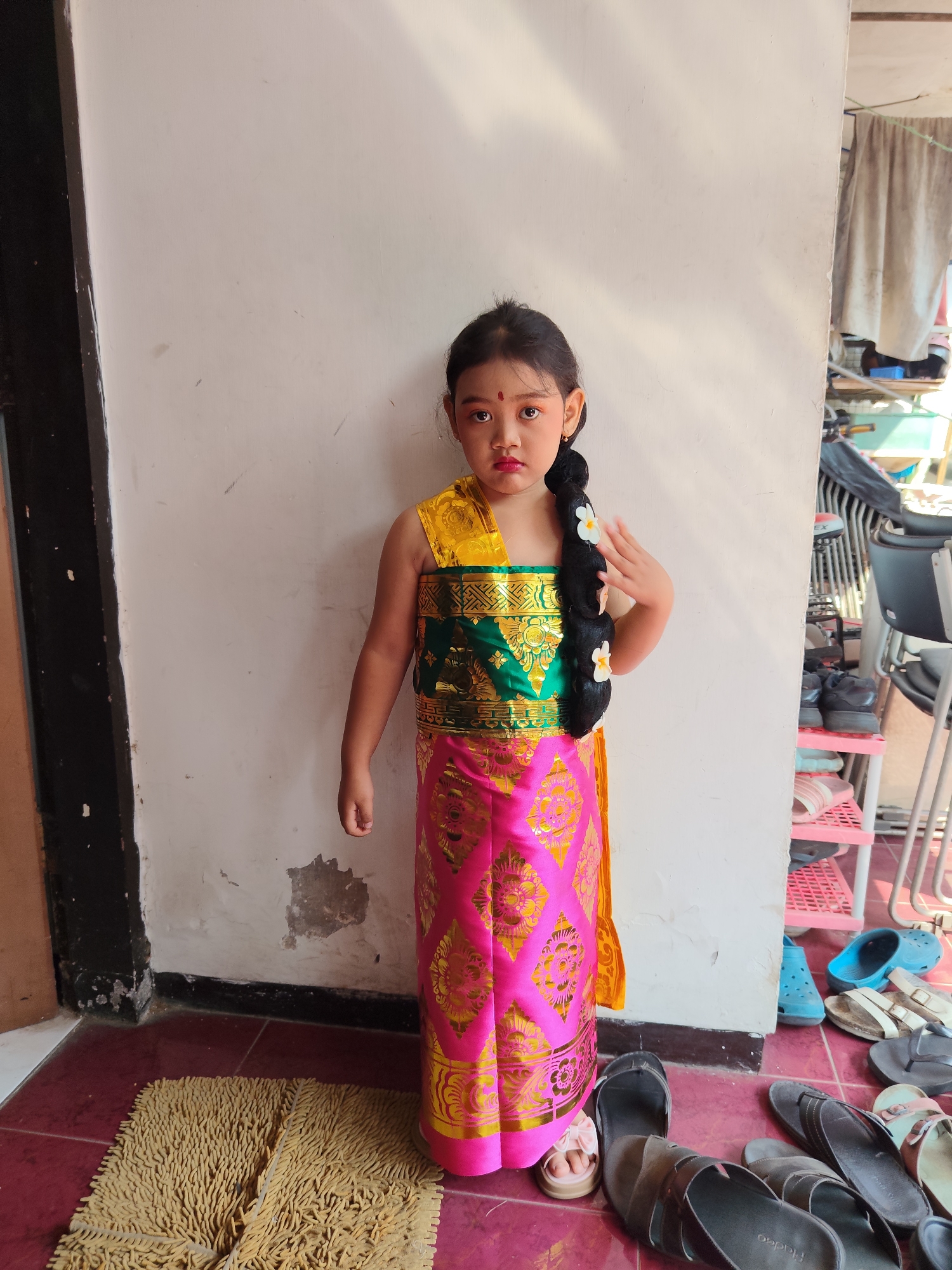A young girl stands in front of a white wall in what appears to be an outdoor setting. She is wearing a traditional Balinese dress, which is a vibrant pink with gold intricate patterns. The dress has a green band across the chest with gold embroidery. She has a long black braid adorned with white flowers. Her makeup is noticeable, with red lipstick and prominent eye makeup. A small red bindi is on her forehead. Her left hand is raised near her braid. In the foreground, a woven mat is visible on the floor. To the right of the girl, there are shelves filled with shoes, and beyond that, what looks like a glimpse of the exterior of a building with laundry hanging. The area appears to be a residential area in the city of Malang, Indonesia, likely a home.