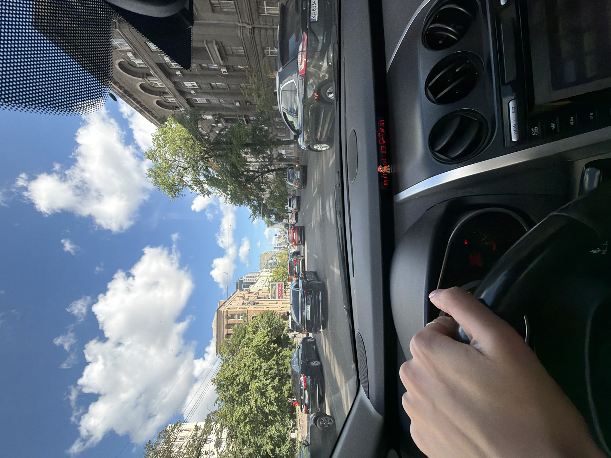The image shows a first-person perspective from inside a car, driving on a city street in Kyiv, Ukraine. The driver's left hand is visible on the steering wheel. The street is lined with trees and multi-story buildings, characteristic of an urban environment. Several cars are on the road, indicating moderate traffic. The sky is blue with scattered white clouds, suggesting a clear, sunny day. The dashboard of the car is visible, with air vents and a display screen. The overall scene depicts a typical urban commute in Kyiv.