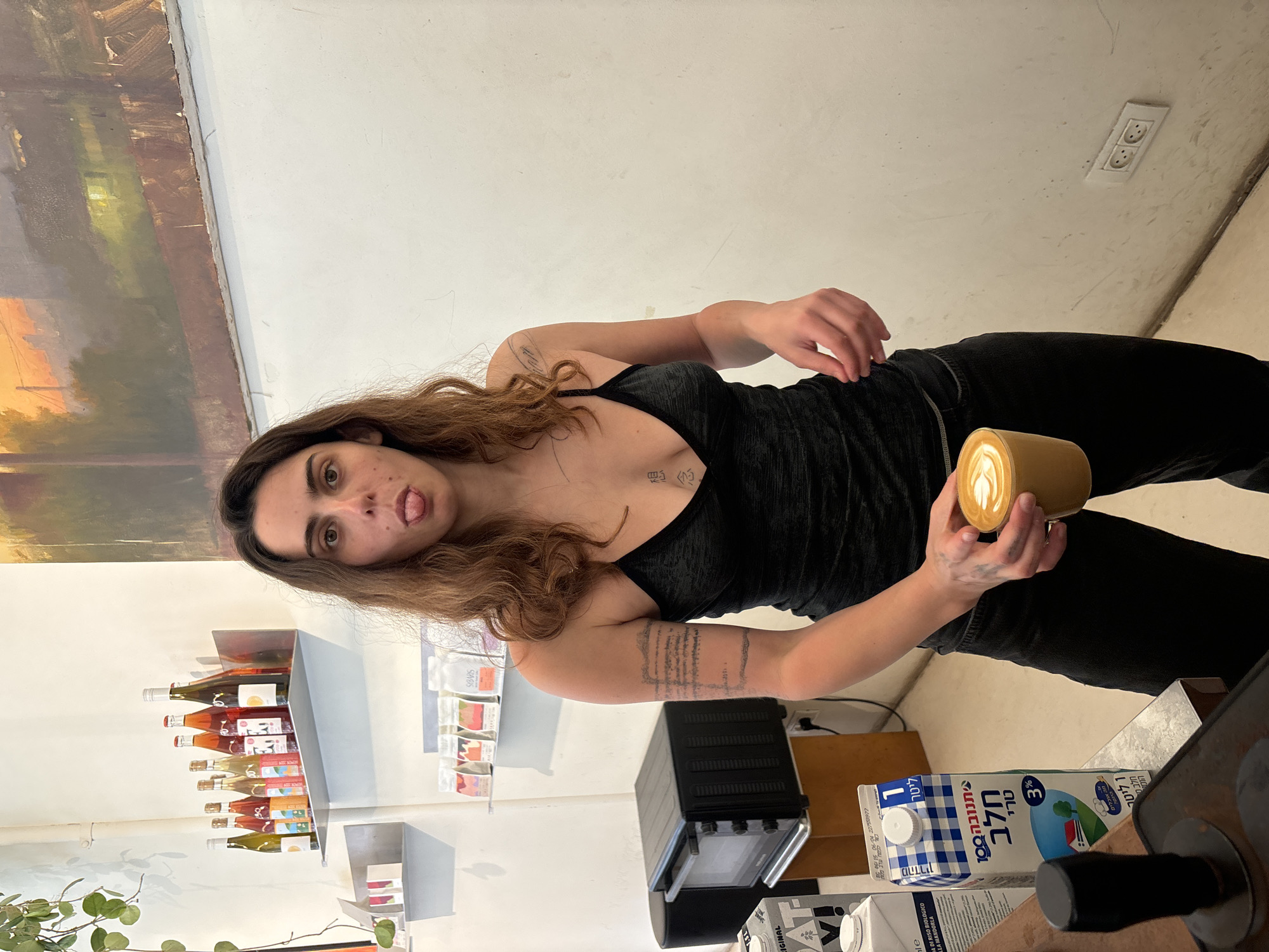 A young woman with brown hair and tattoos is holding a cup of coffee with latte art in a room in Tel Aviv, Israel. She is wearing a black top and pants, and sticking her tongue out. Behind her, there are shelves with wine bottles, food products, and a toaster oven. There is a carton of milk on the counter. A painting is visible on the wall. An electrical outlet is visible.