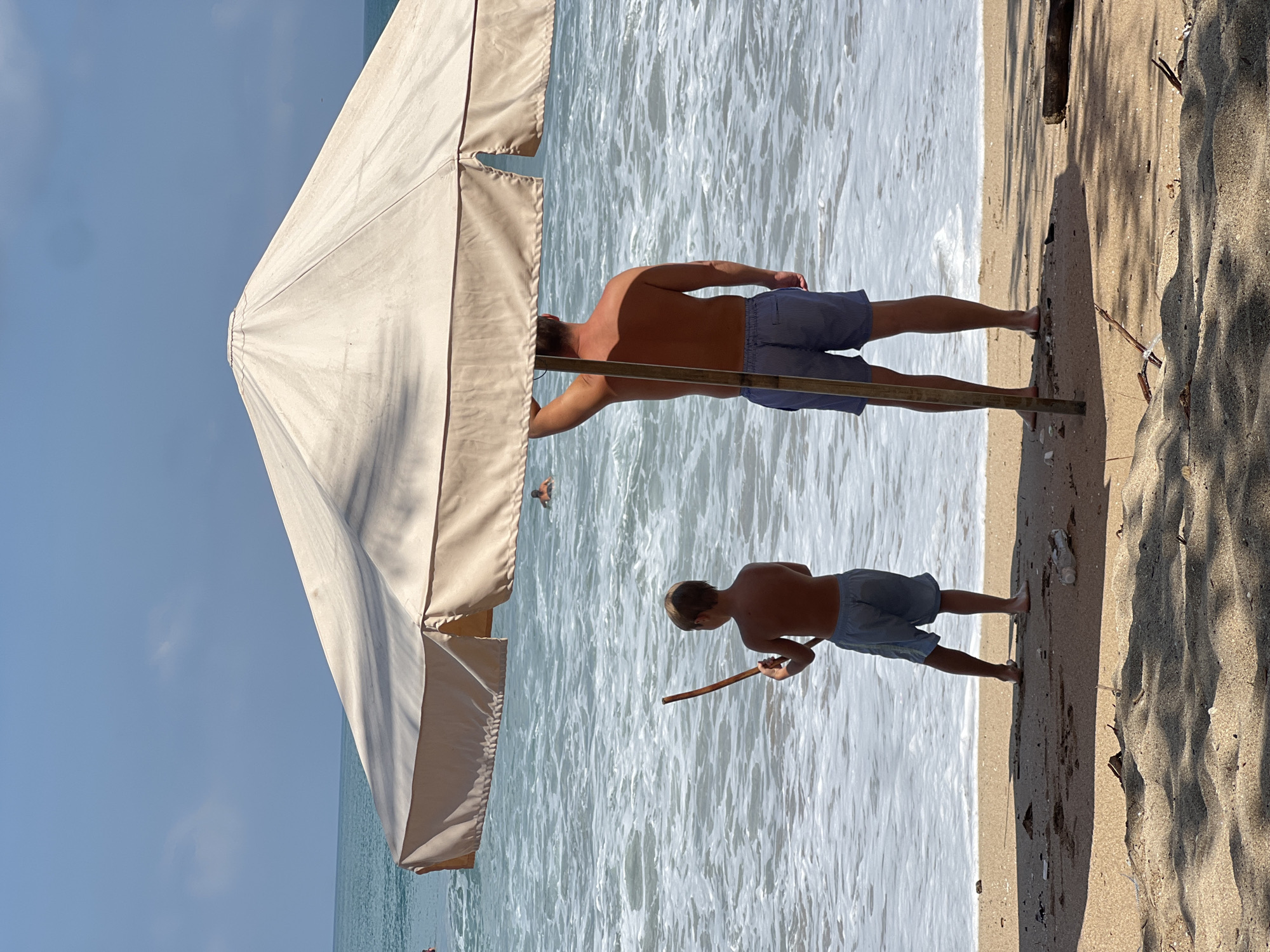 An eye-level, medium shot captures a boy and a man standing on the shore of Kuta Beach, Indonesia, under a large beach umbrella. The boy, on the left, faces the ocean, holding a stick. He wears light blue shorts. The man, to the right, faces away from the camera, also looking out at the water. He is shirtless and wears blue and white striped shorts. He holds his left arm up, seemingly adjusting the umbrella. The ocean waves are foamy and white, with a clear blue sky overhead. The sand in the foreground is light brown, with some shadows from the umbrella and palm trees. The general atmosphere suggests a sunny day at the beach.